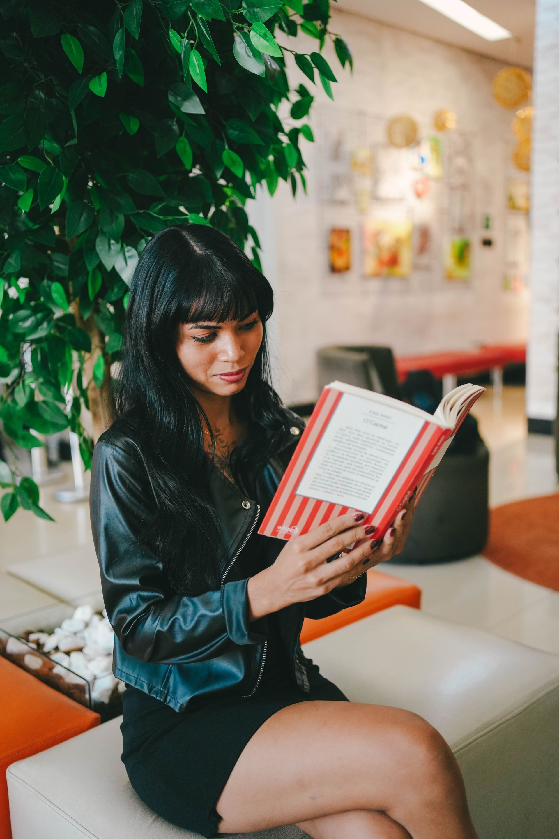 Uma mulher de jaqueta preta lê um livro, sentada perto de uma planta frondosa em um interior moderno com detalhes em laranja.