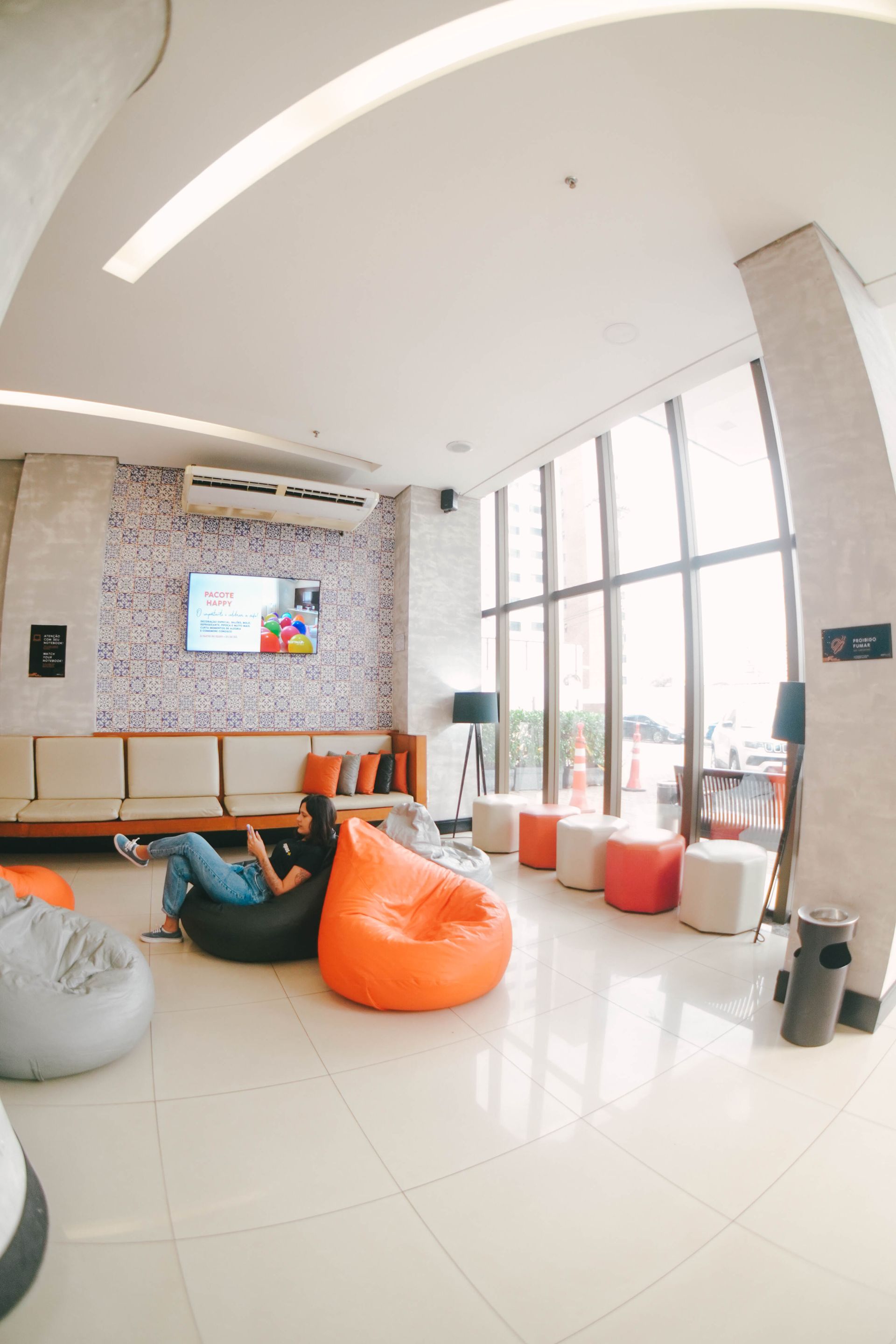 Área de estar com pufes, sofás, TV e grandes janelas. Uma pessoa relaxa em um pufe.