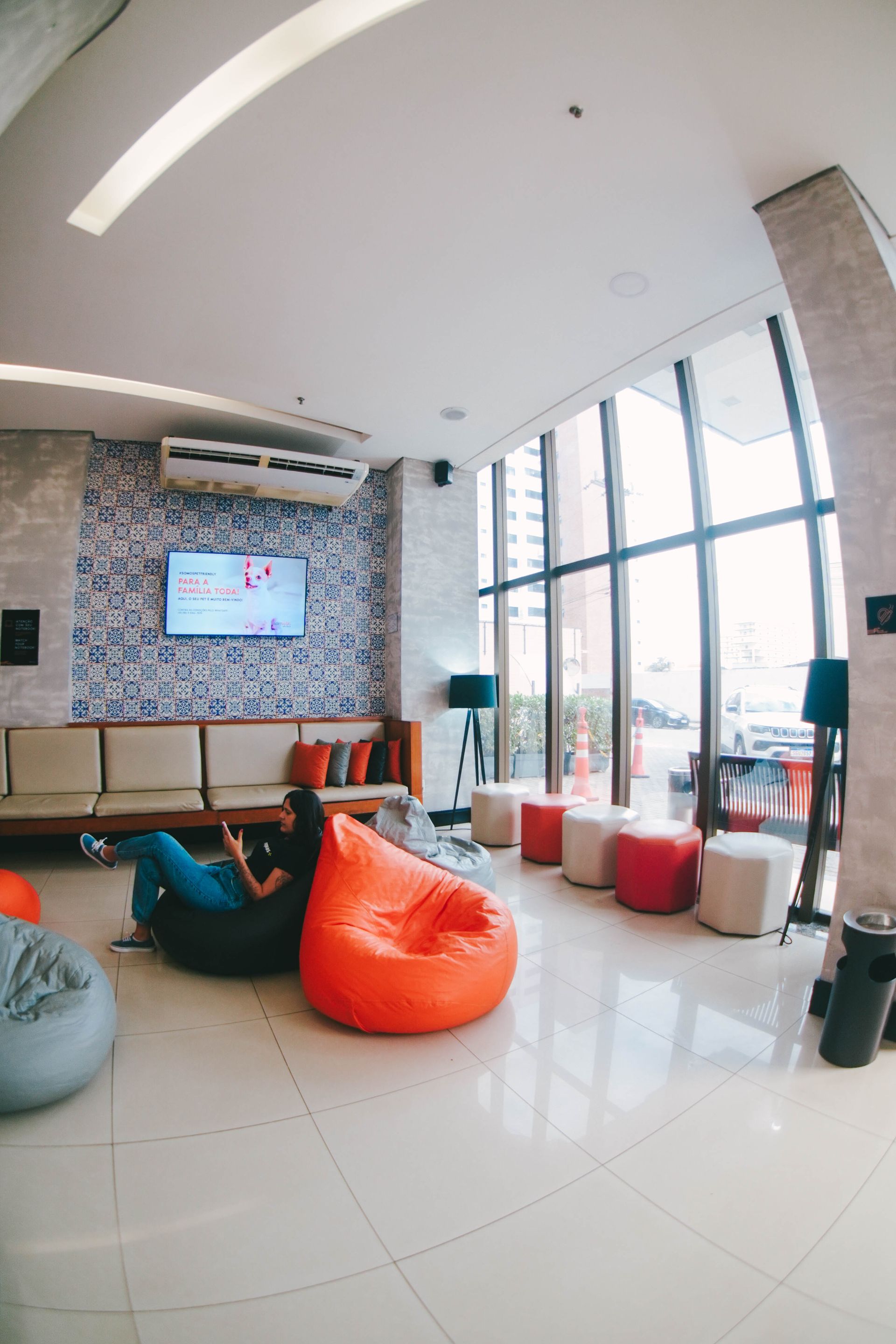 Área de estar com pufes e janela com vista para a rua. Uma pessoa está reclinada em um pufe.