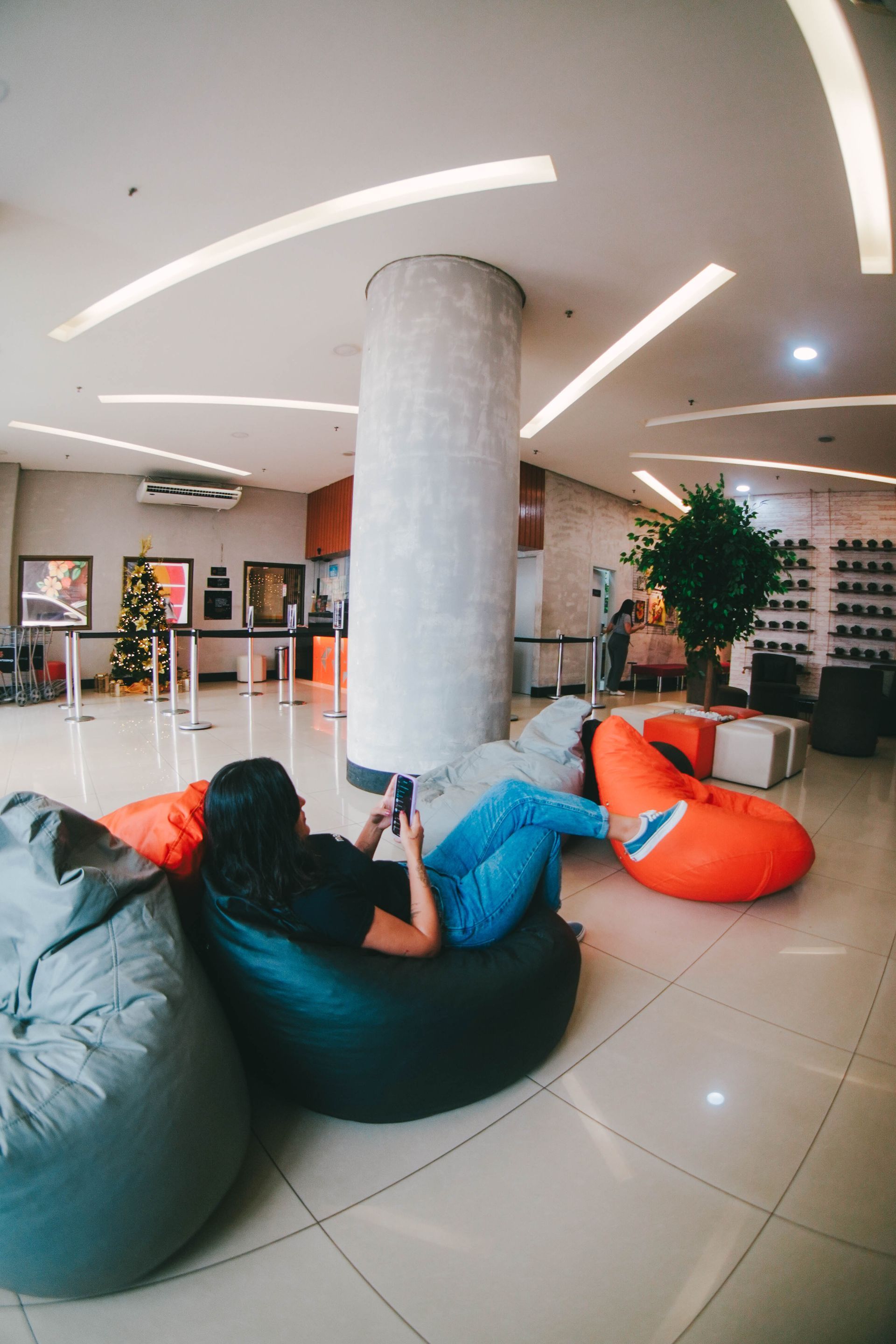 Pessoa sentada em uma poltrona de saco de feijão em um saguão, usando um telefone. Outras poltronas de saco de feijão e um grande pilar também são visíveis.