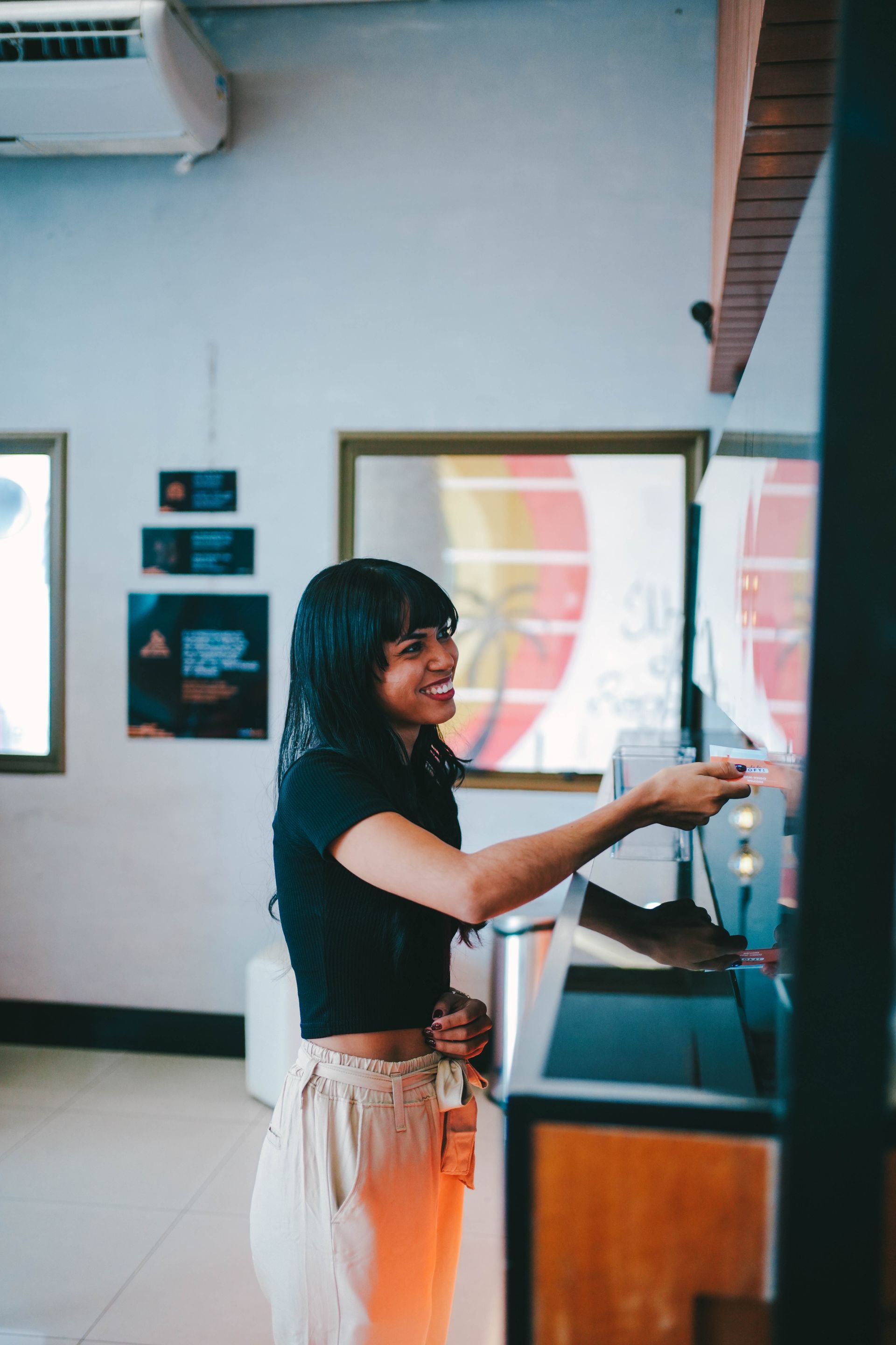 Mulher sorridente estende a mão em direção a uma máquina em um ambiente bem iluminado. Blusa preta, calça bege.