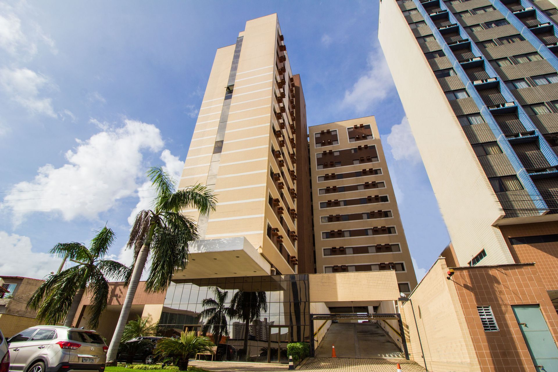Prédio alto bege com entrada de vidro, carro e palmeiras sob um céu claro.