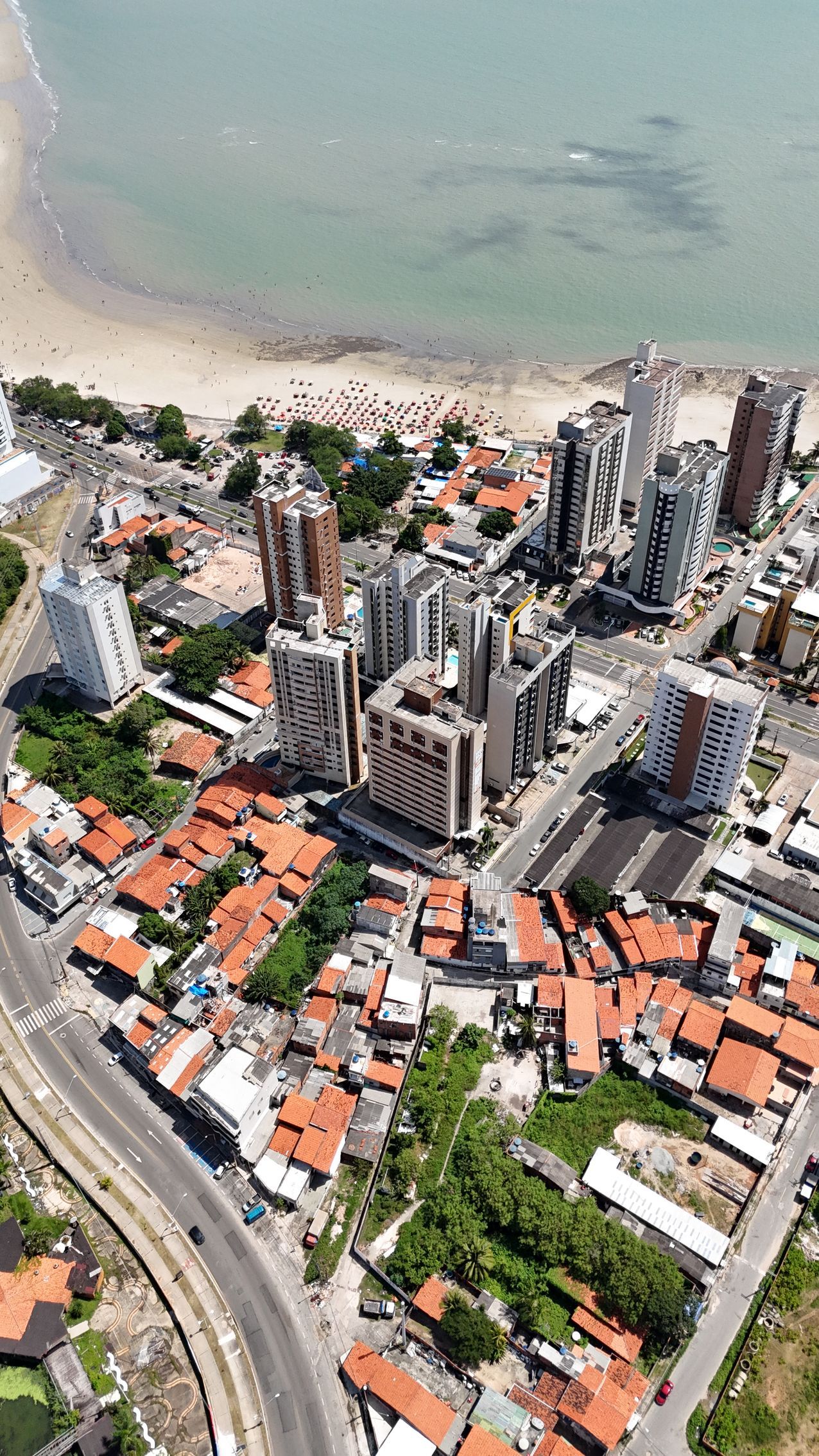 Vista aérea de uma cidade costeira com edifícios altos, praia e áreas residenciais com telhados vermelhos.