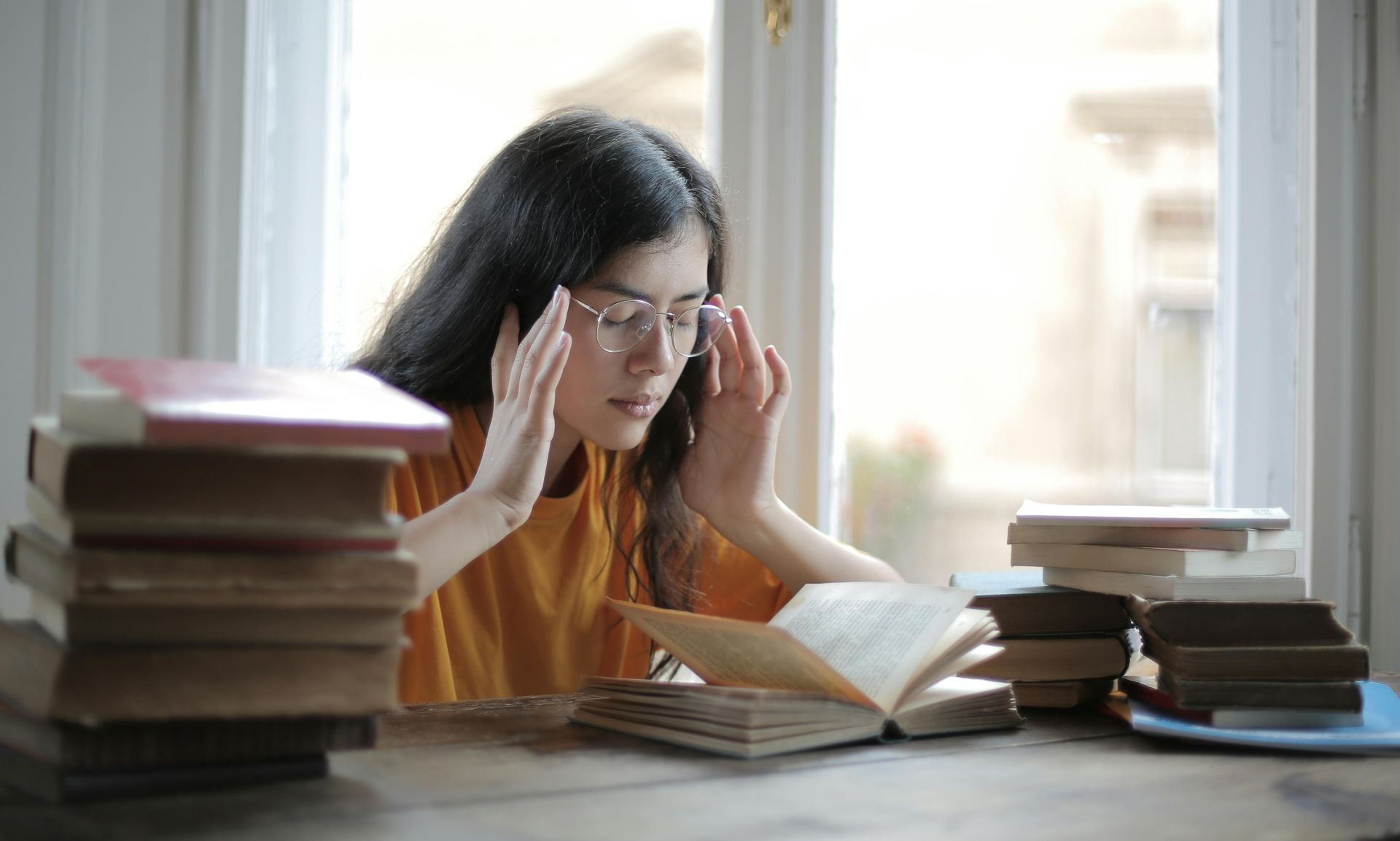 A woman is sitting at a table with a stack of books and reading a book.