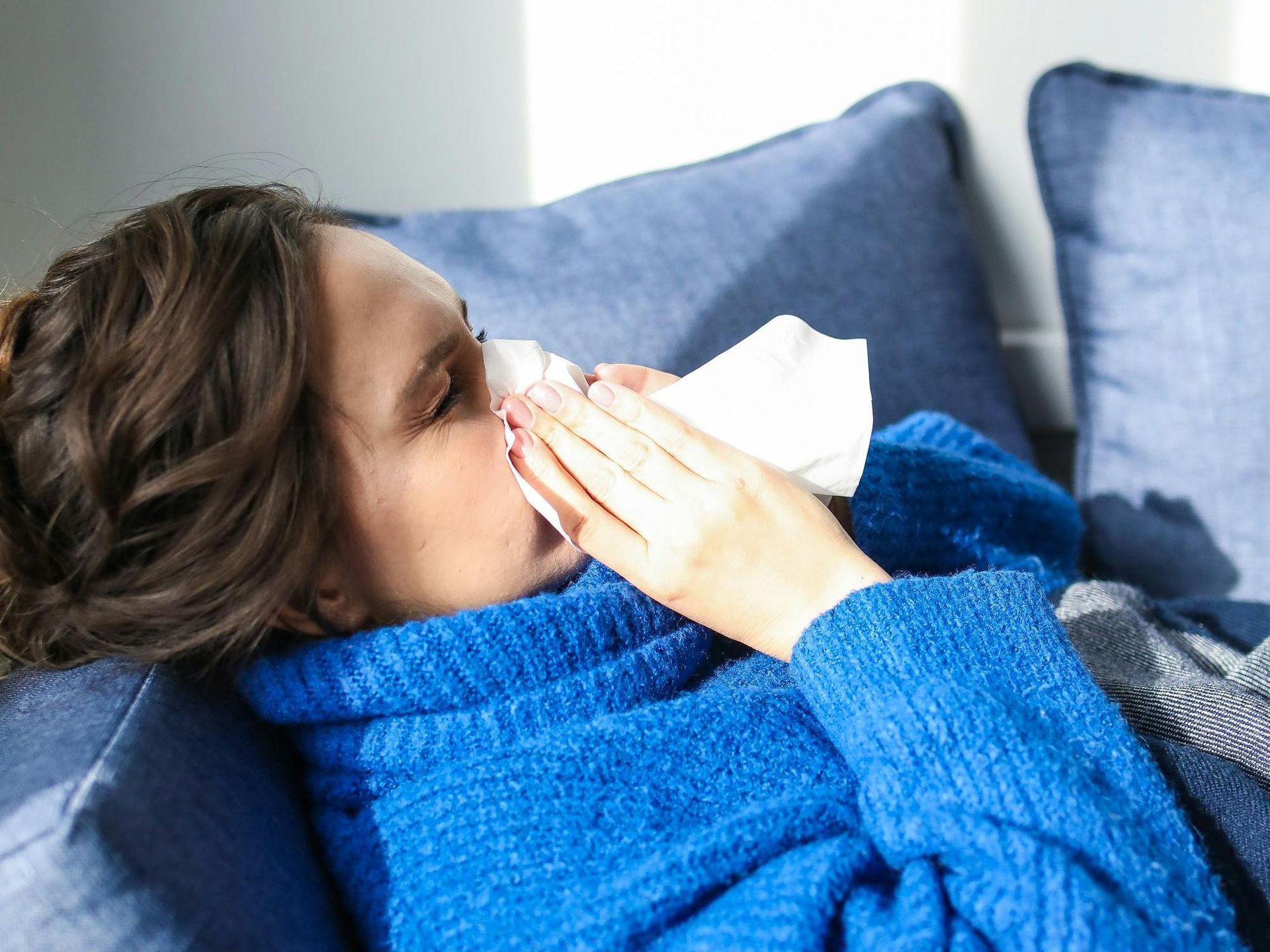 A woman is laying on a couch blowing her nose with a napkin.
