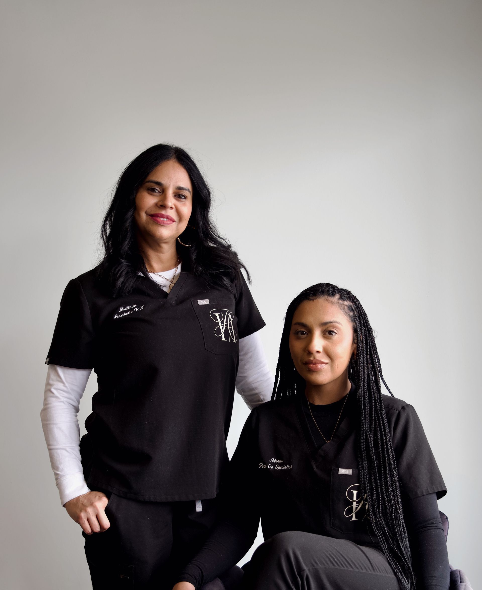 Two women in scrubs are posing for a picture while one is sitting in a chair.