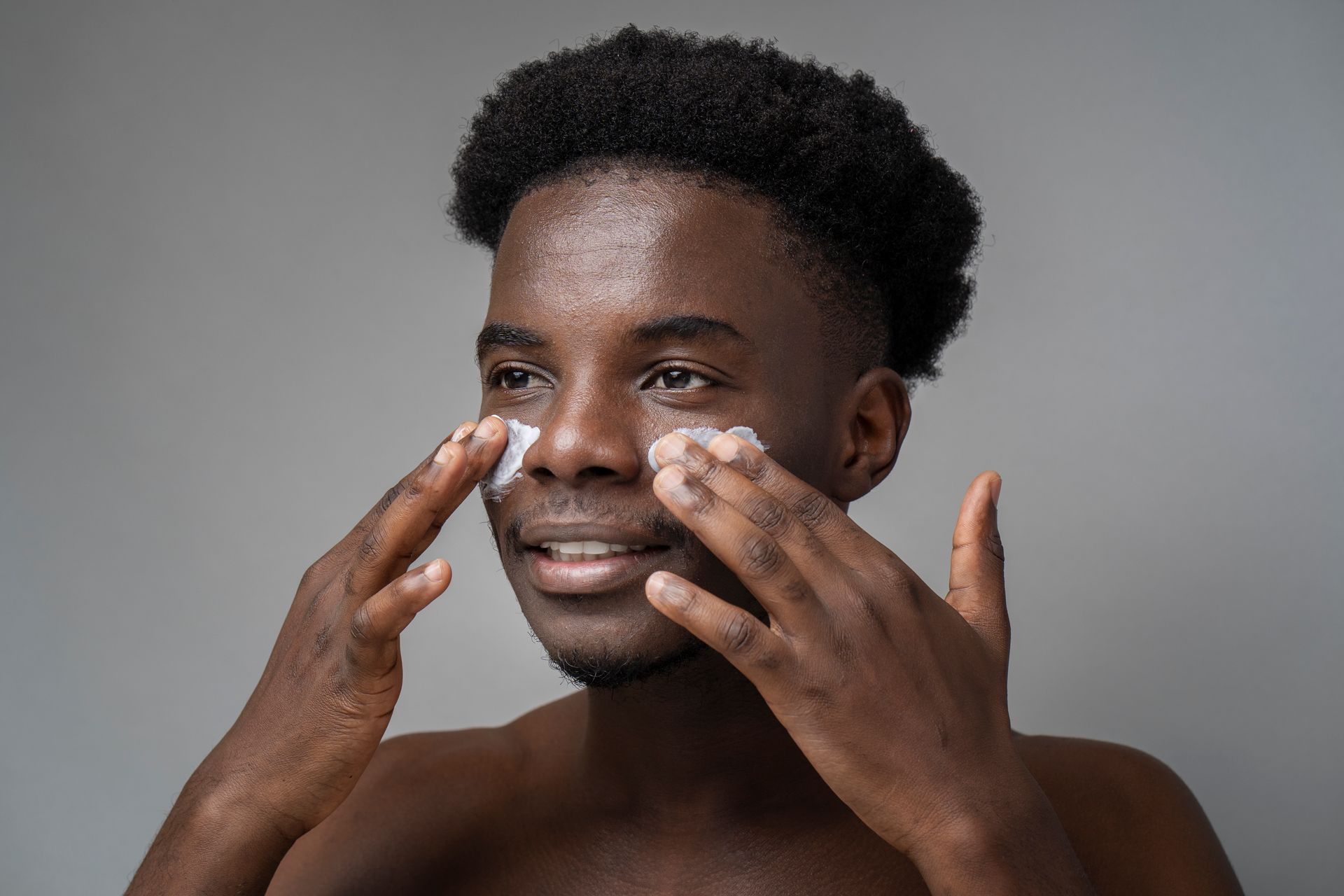 A shirtless man is applying lotion to his face.