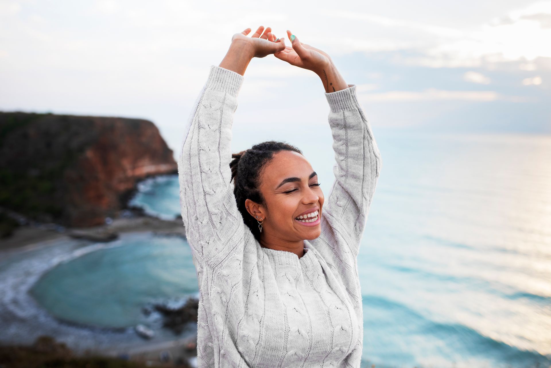 A woman is standing on top of a cliff overlooking the ocean with her arms in the air.