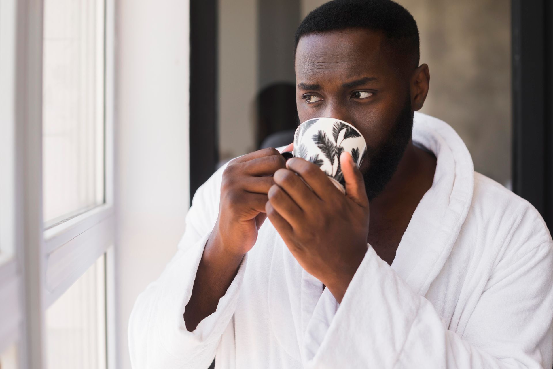 A man in a white robe is drinking a cup of coffee while looking out of a window.