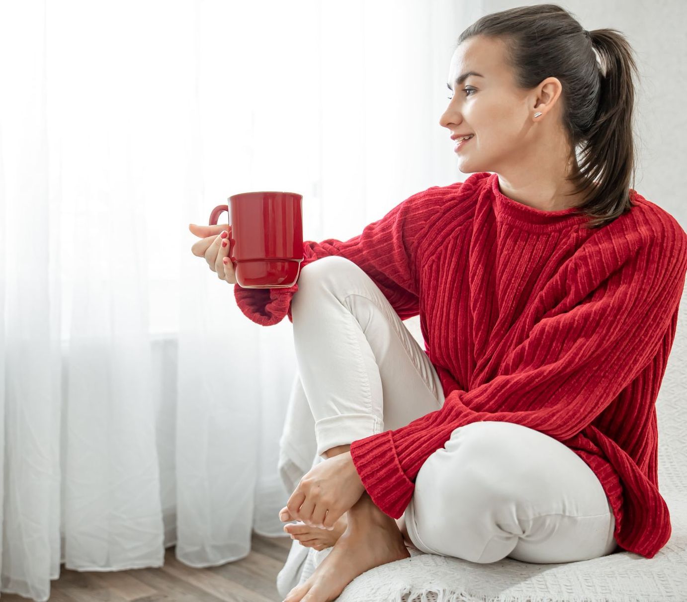A woman in a red sweater is sitting on a window sill holding a cup of coffee.