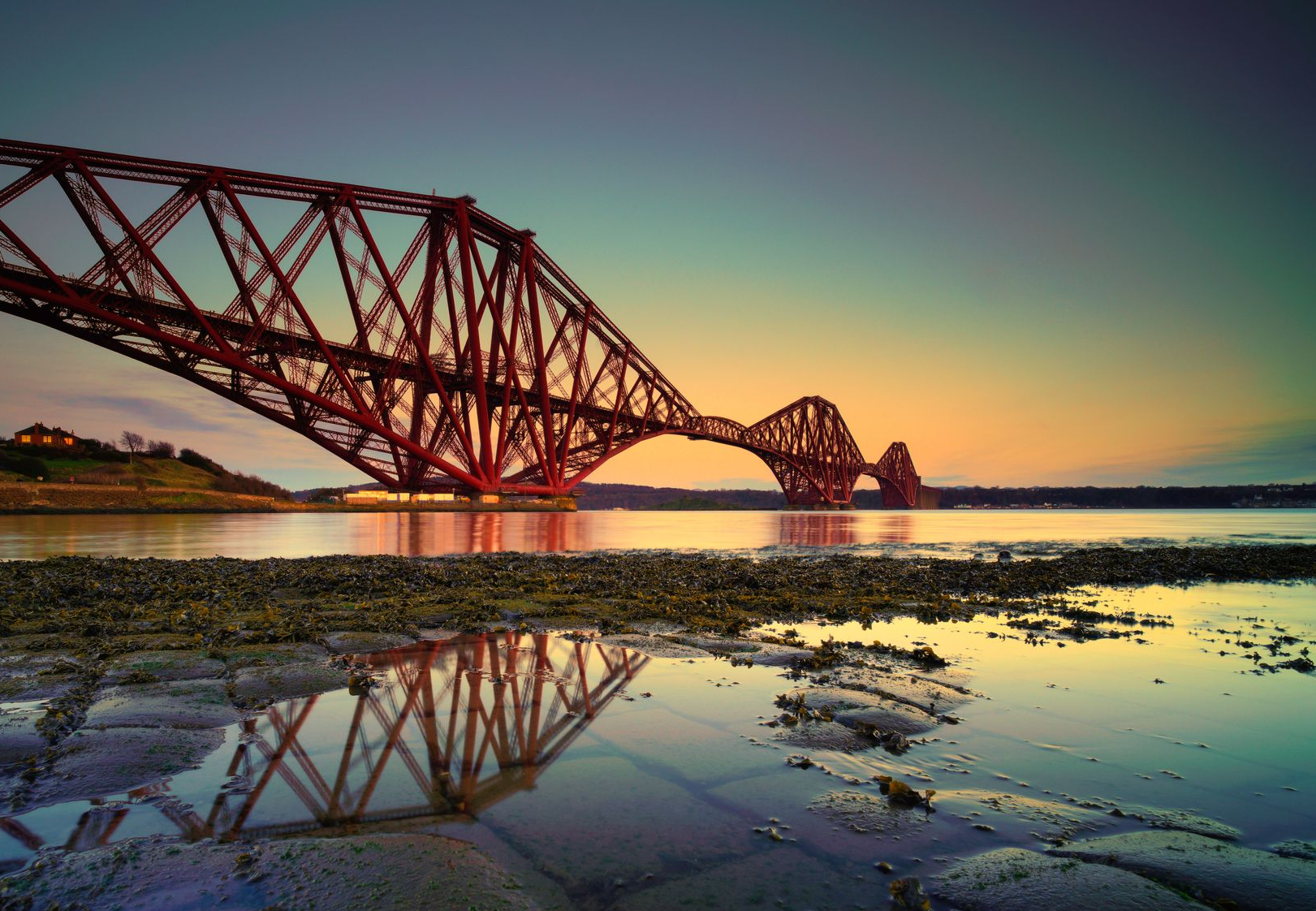 The iconic red Forth Bridge in Scotland silhouetted against a golden sunrise, with its reflection in a tidal pool.
