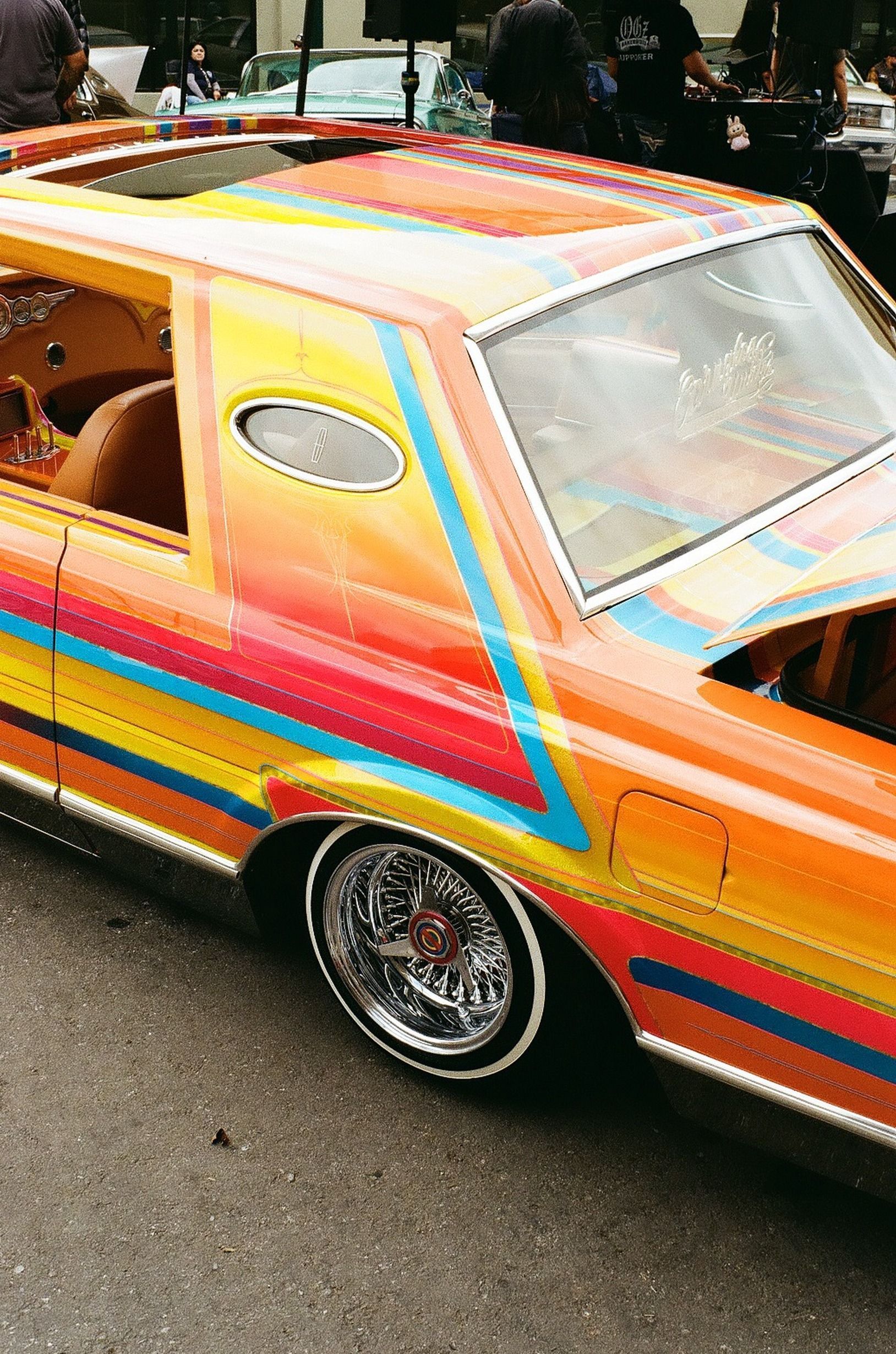 Bright orange lowrider car with rainbow stripes and chrome wheels at an outdoor car show