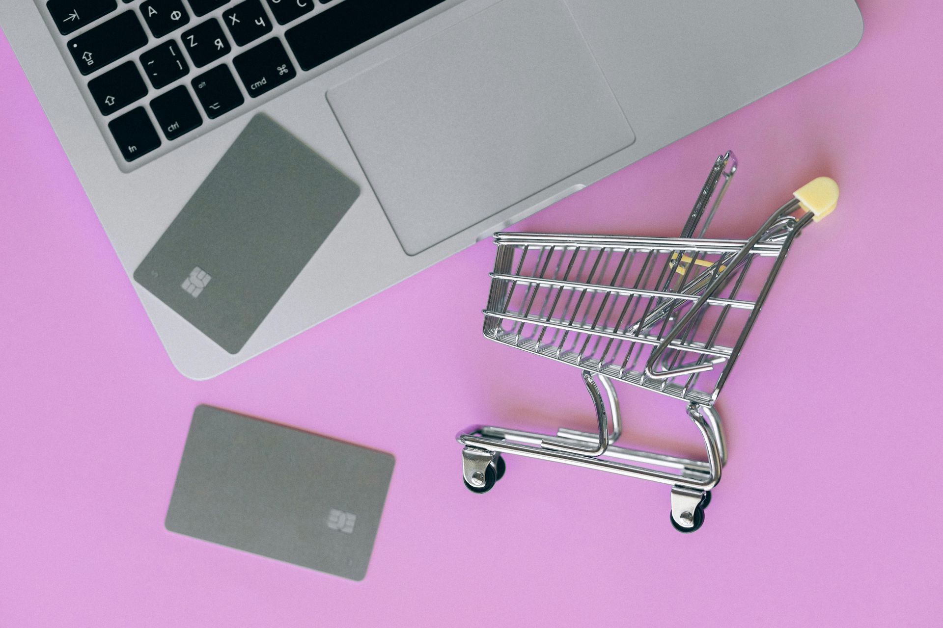 Laptop, shopping cart, and two gray credit cards on a pink surface, suggesting online shopping.