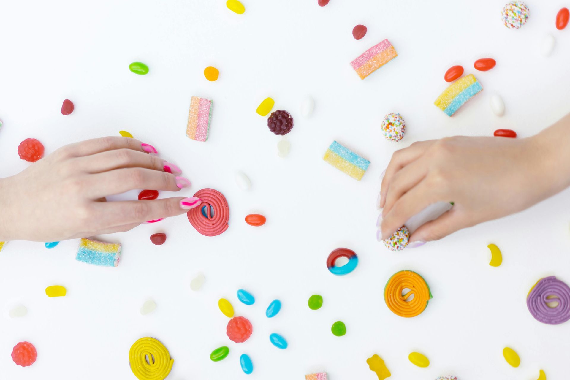 Hands reaching for candy scattered on a white surface; colorful gummies, jelly beans.