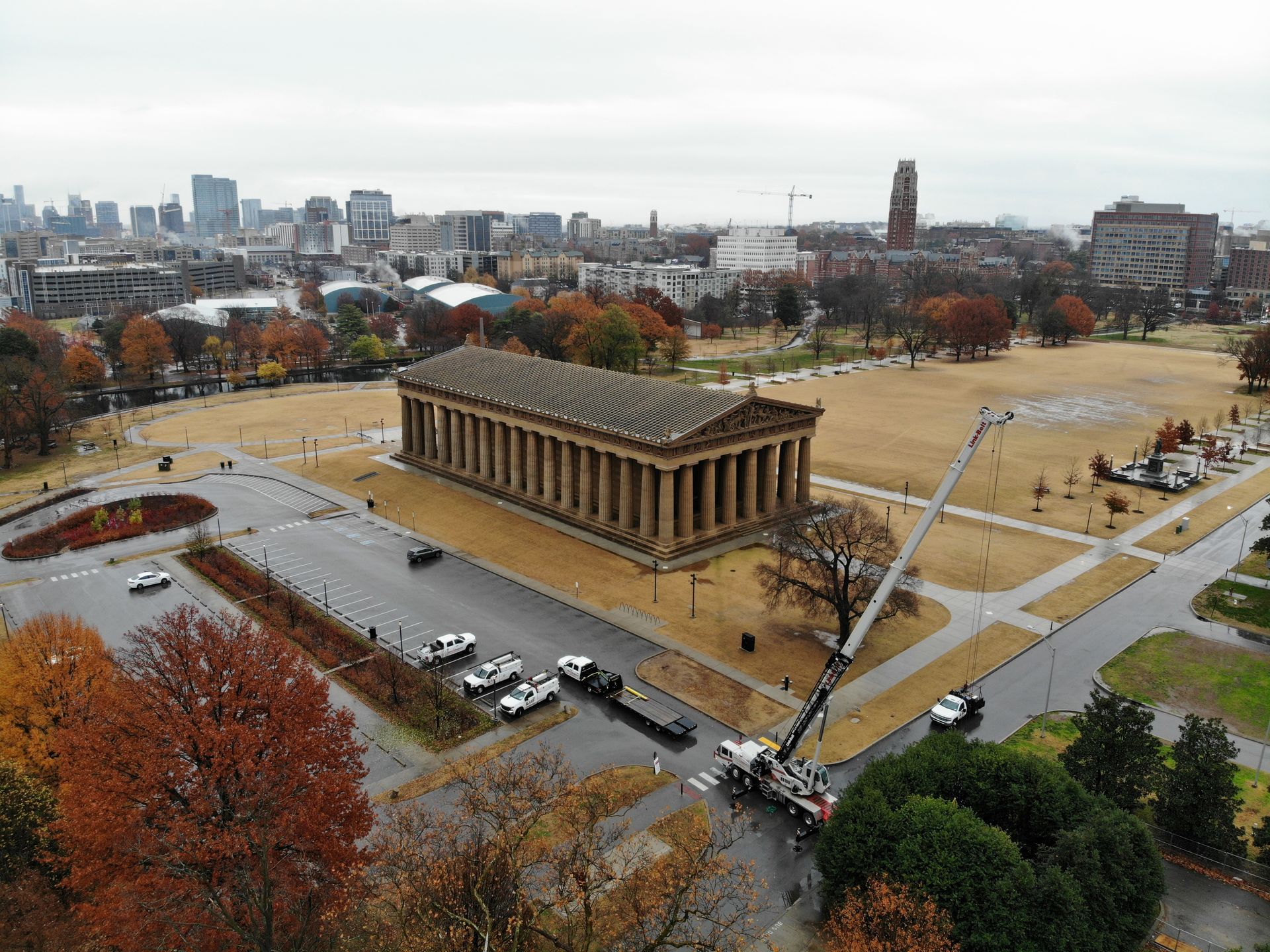 Aerial view of a Greek temple-like structure with a city skyline in the background, surrounded by trees and a parking lot.