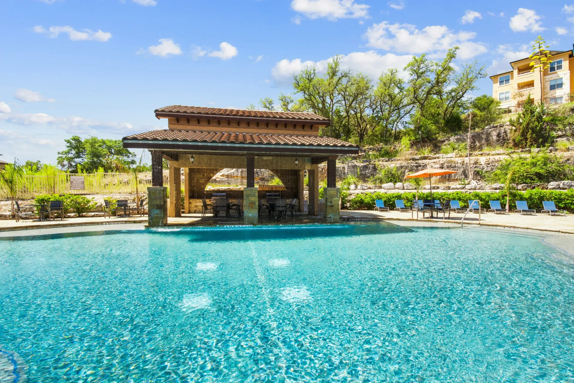 Pool area with lounge chairs, blue water, and stucco buildings under a blue sky.