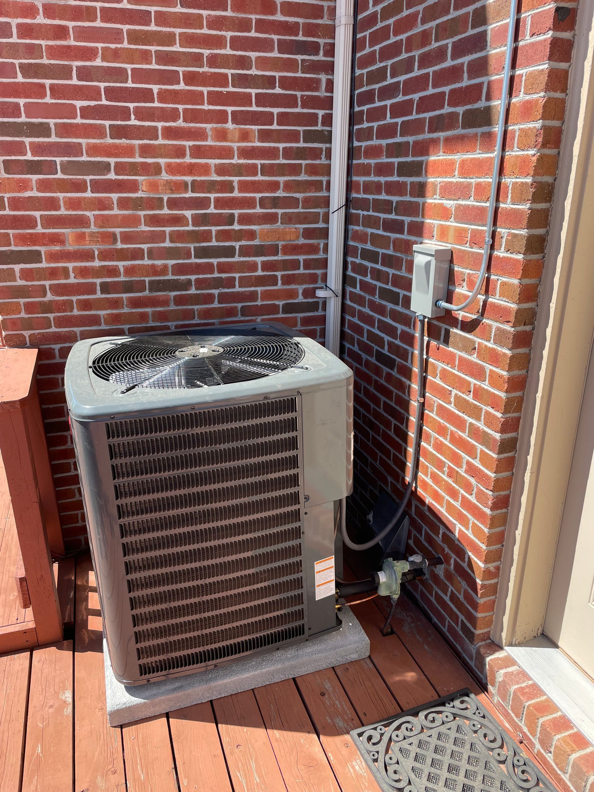 An air conditioner is sitting on a wooden deck next to a brick wall.