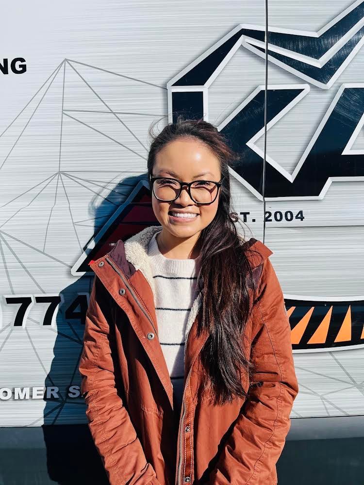 A woman wearing glasses and a brown jacket is standing in front of a van.