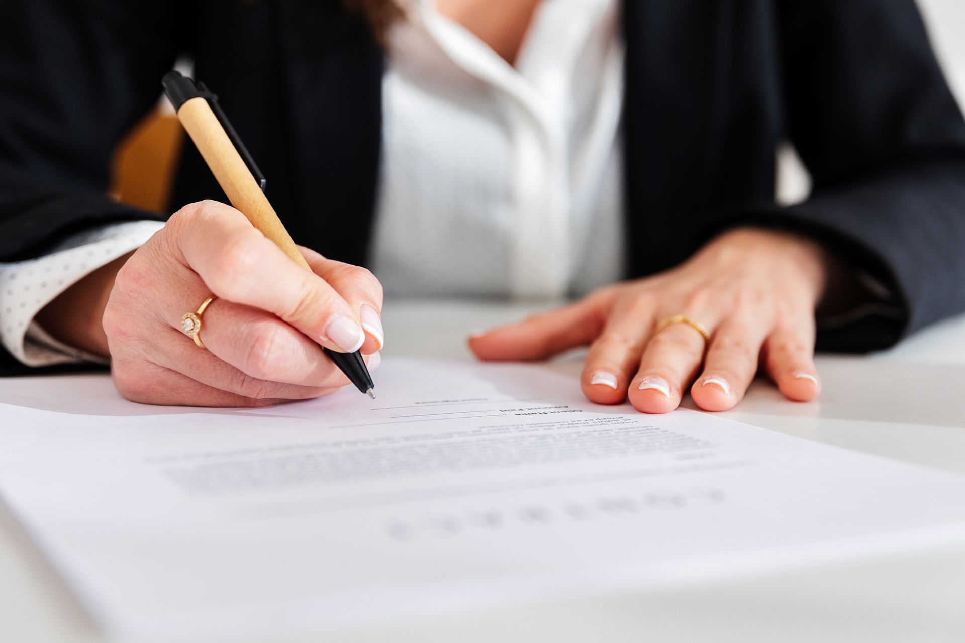 Woman in a black blazer signing a document with a pen, rings on her fingers.