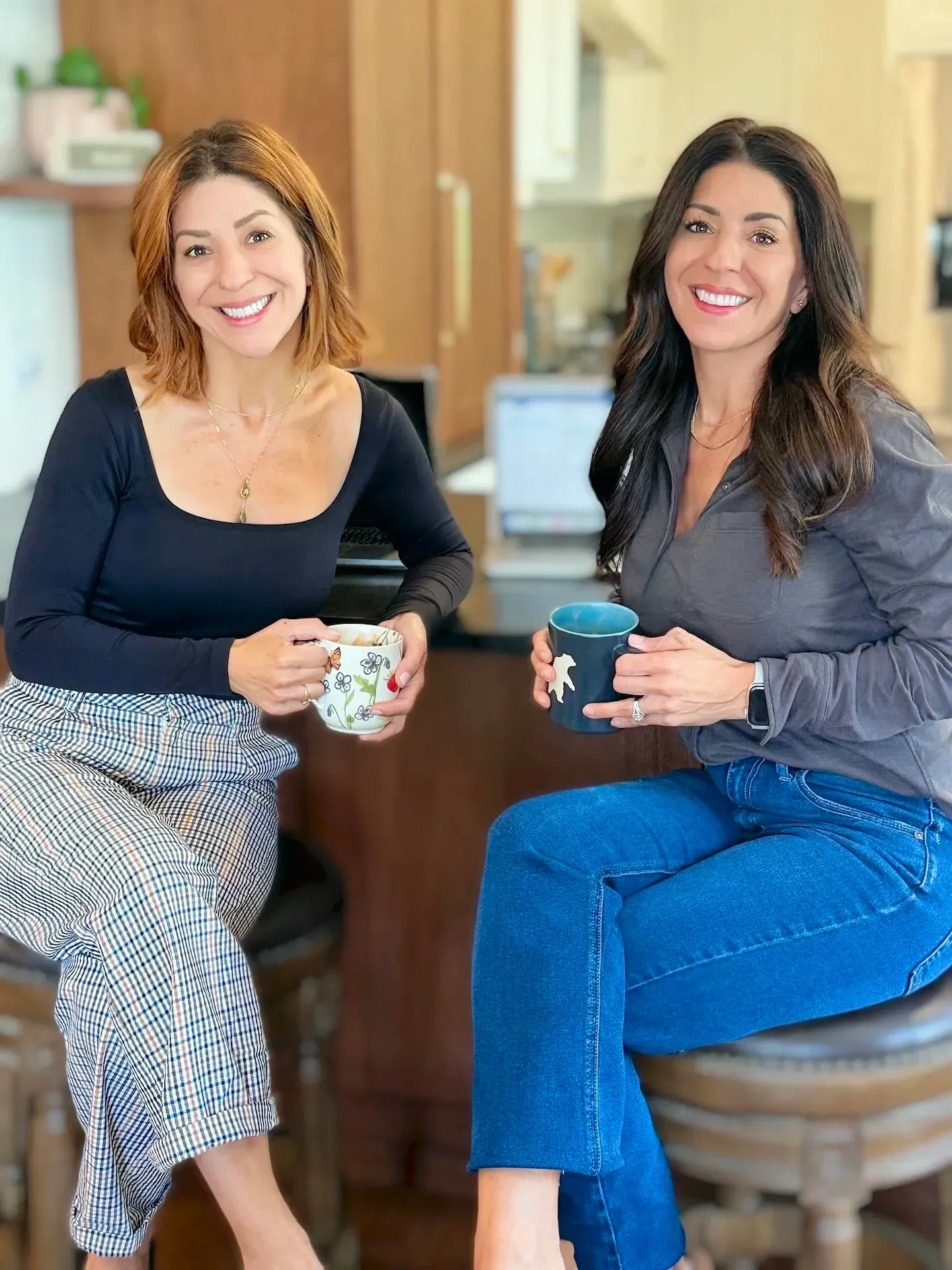Two women, sitting, smiling, holding mugs. Kitchen setting. One wears black top, patterned pants; the other wears a grey top, jeans.