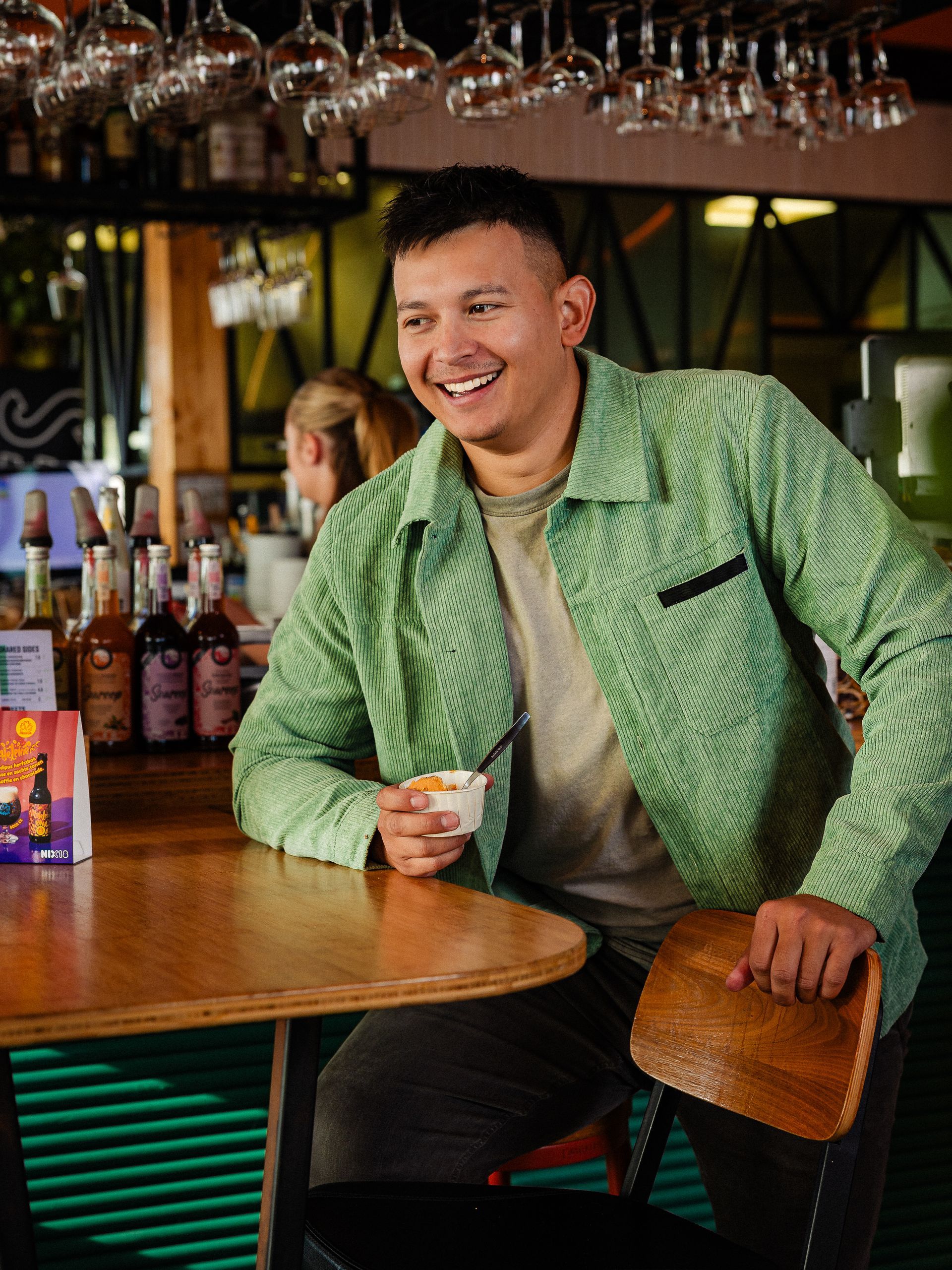 Een man in een groene jas glimlacht terwijl hij een beker vasthoudt aan de bar.