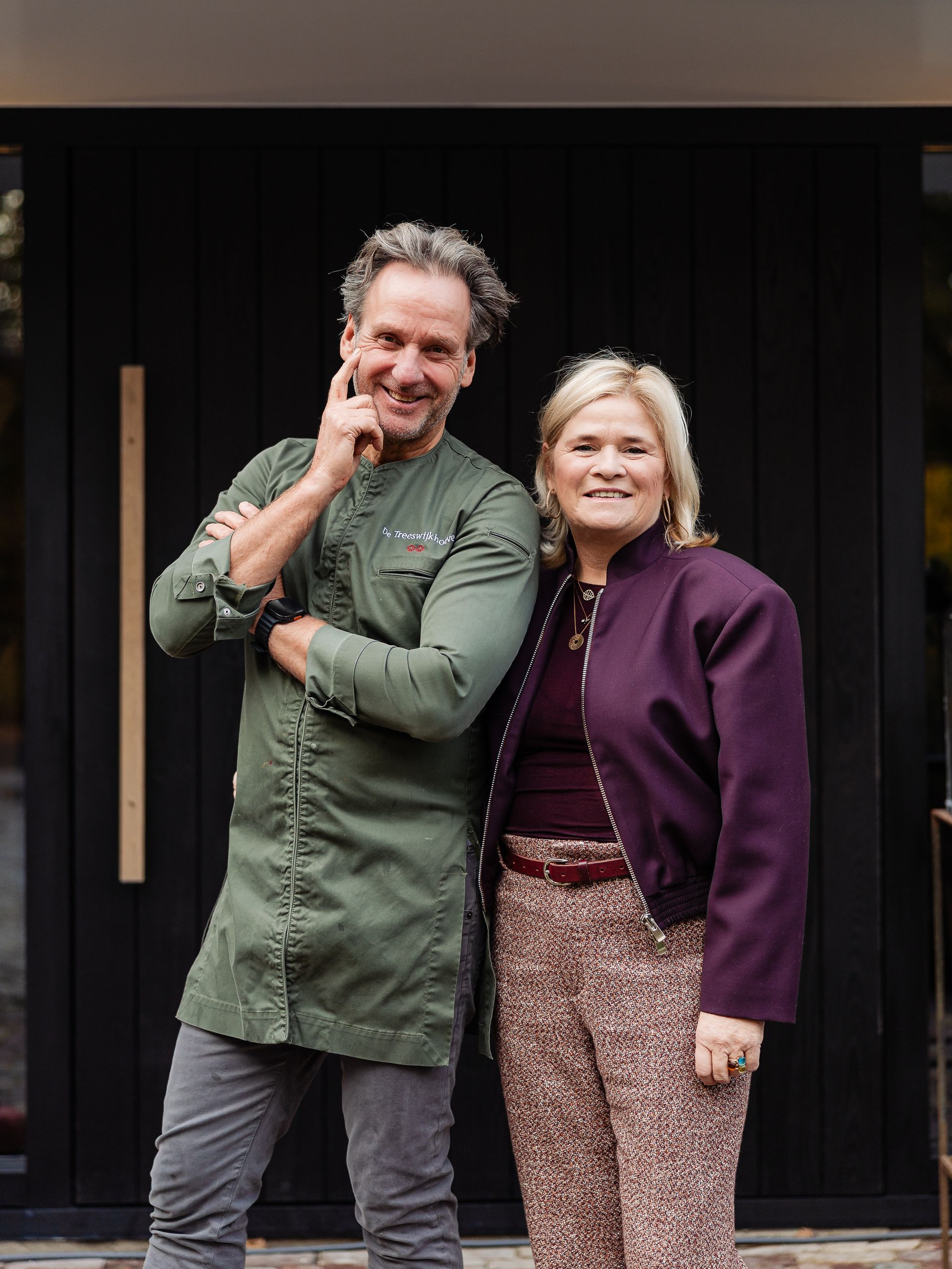 De chef-kok glimlacht, terwijl hij een donker dessert in een witte kom vasthoudt, gekleed in een witte koksjas en een zwart schort.