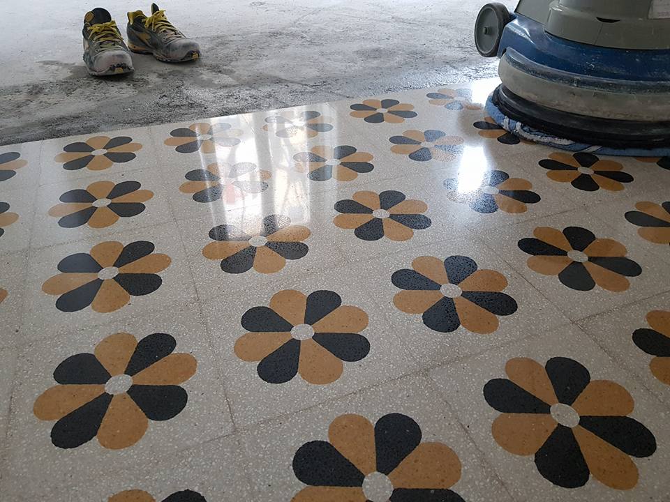 Pavimento in piastrelle lucide con motivi floreali neri e dorati. Sono visibili una lucidatrice e degli stivali da lavoro.