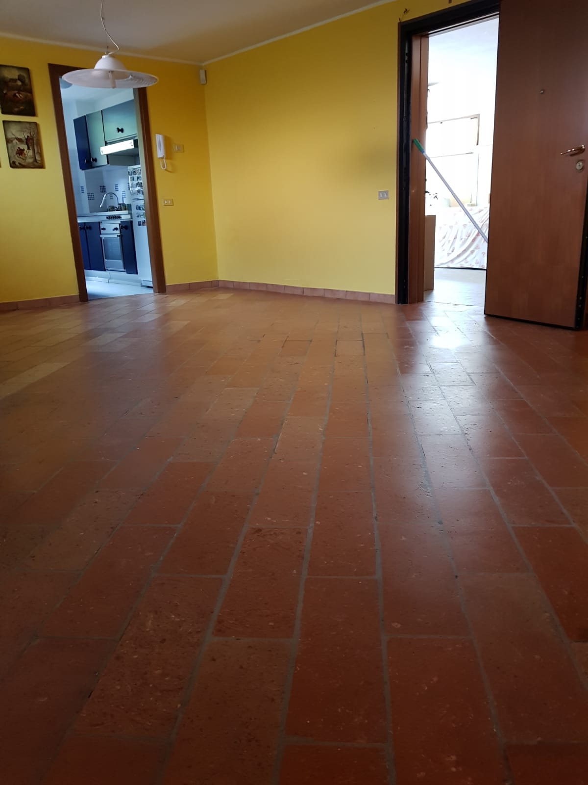 Stanza vuota con pavimento in cotto, pareti gialle e porte aperte sulla cucina e sull'esterno.