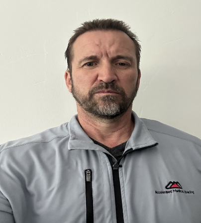 Man in a light gray jacket with a logo, standing against a plain wall, looking serious.
