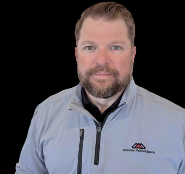 Man wearing a light gray jacket with a logo, standing against a black background