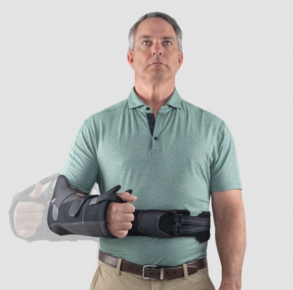 Man wearing a black arm brace and sling, holding his left forearm against his chest on a white background