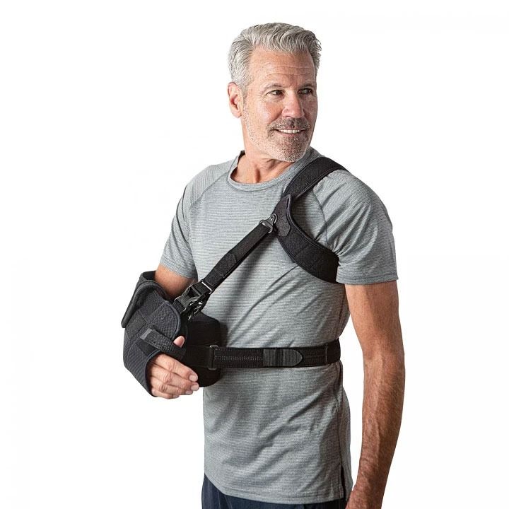 Man in a gray T-shirt wearing a black arm sling and shoulder brace on a white background