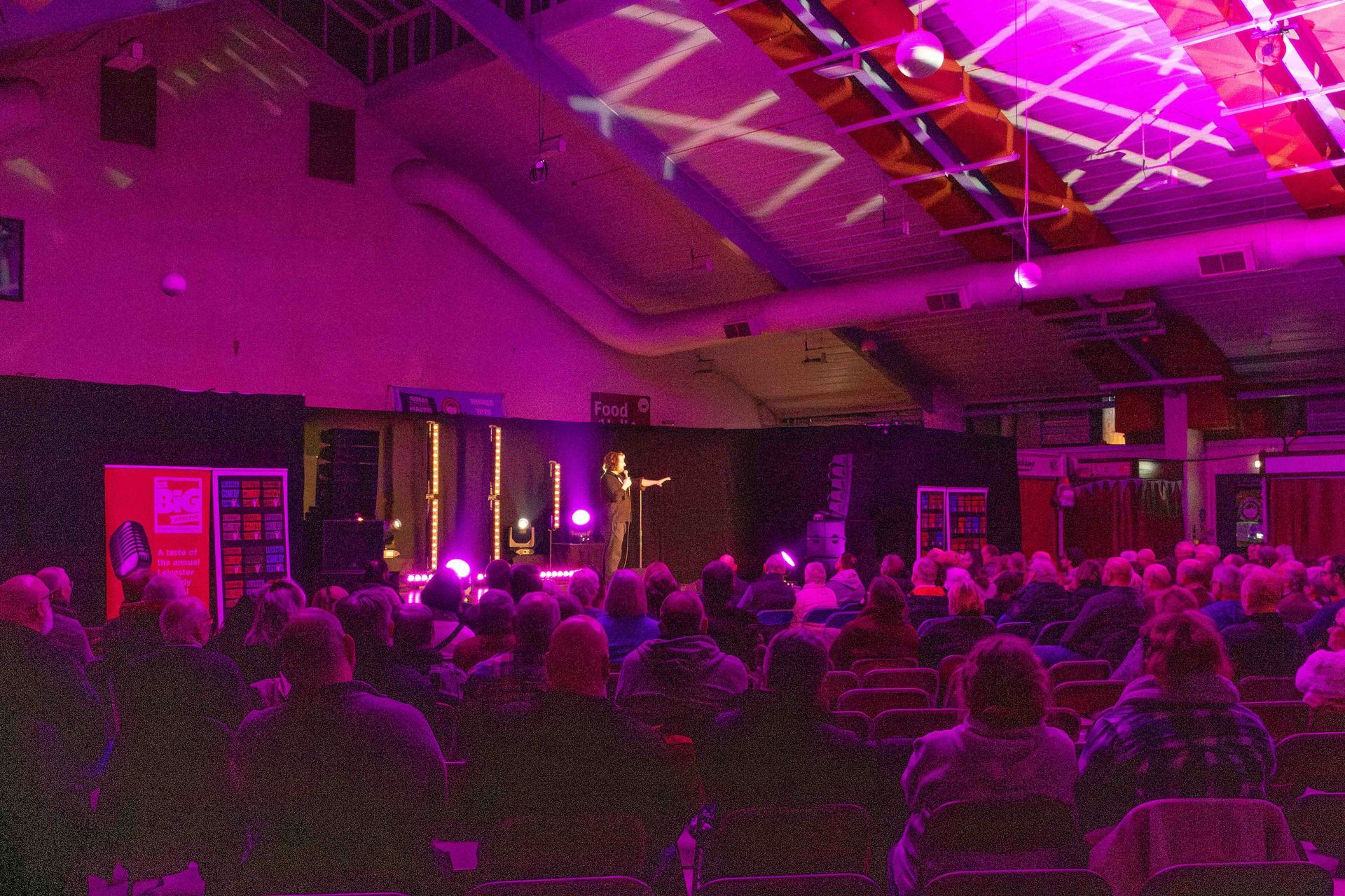 Audience watches a performance on a stage lit with purple lights inside a large hall.