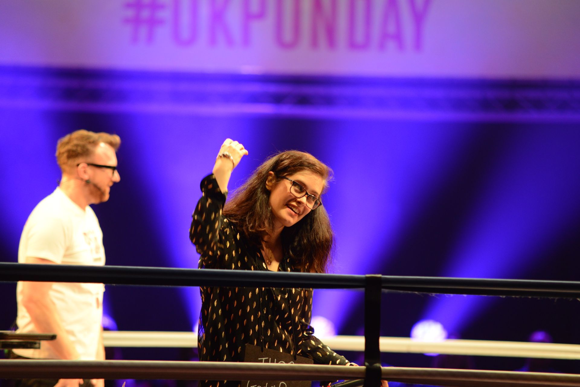 Woman in glasses raises arm victoriously in a boxing ring, #UKPunday sign in background. Man looks on.