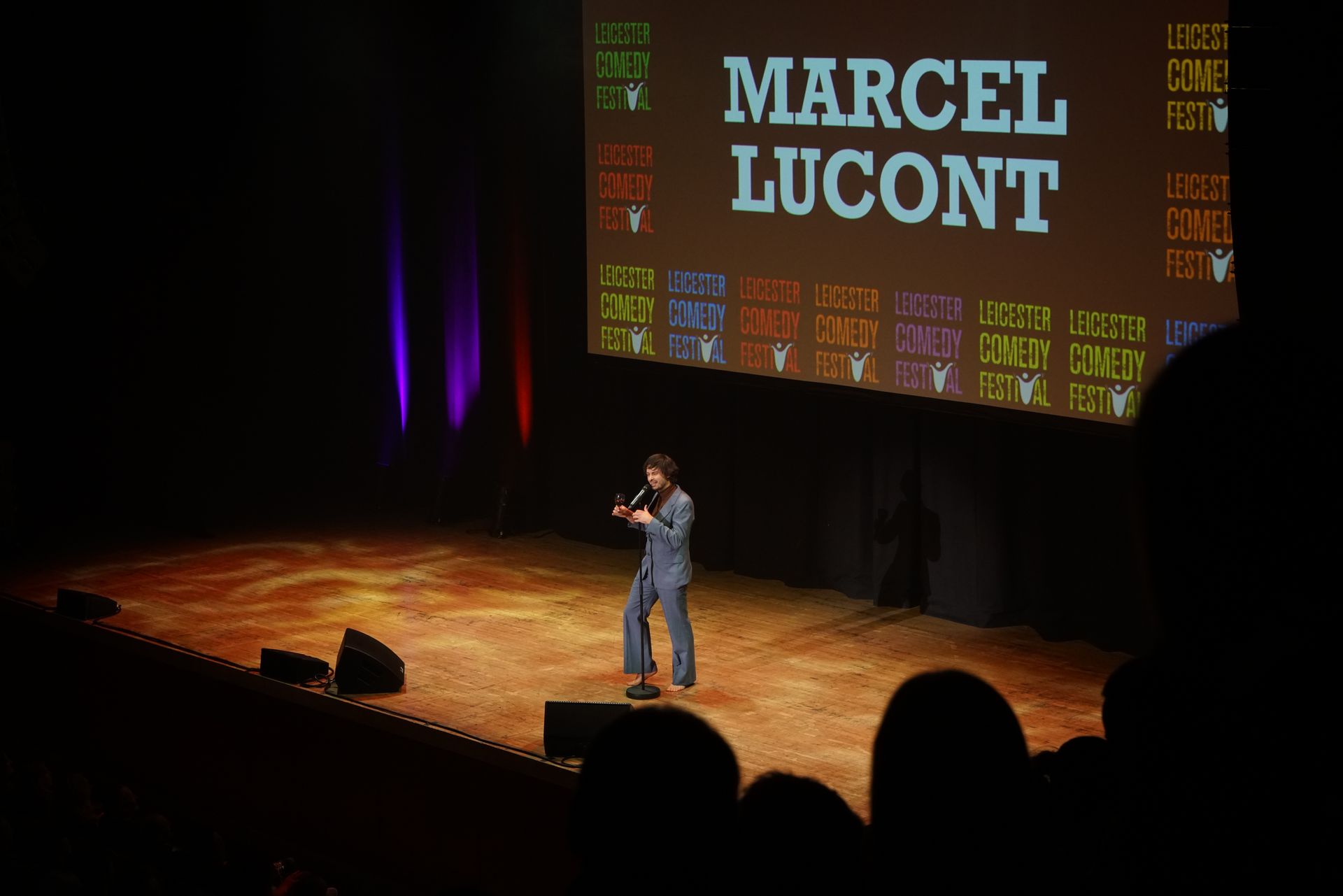 Marcel Lucont performing stand-up on stage. Stage lights and audience visible. Festival banner in background.