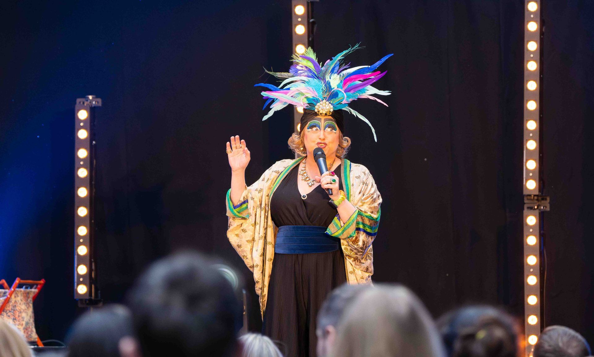 Drag performer on stage, wearing a feathered headdress, speaking into a microphone.