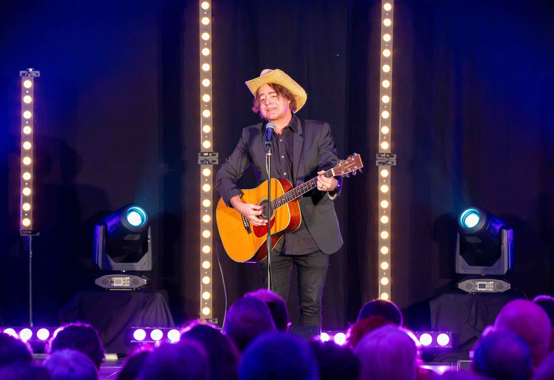A performer on stage with a guitar, wearing a hat, performing in front of a crowd.