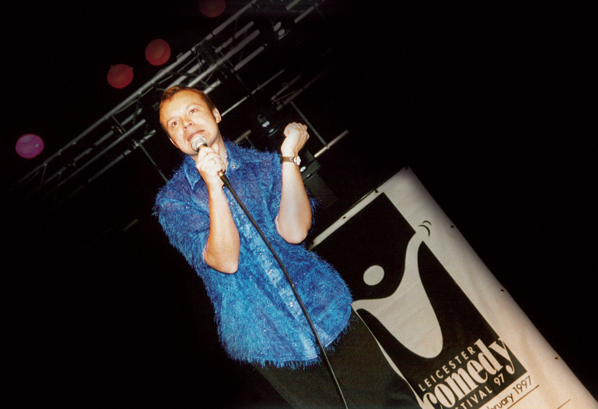 Comedian on stage at the Gloucester Comedy Festival, holding microphone, dark setting, blue shirt.