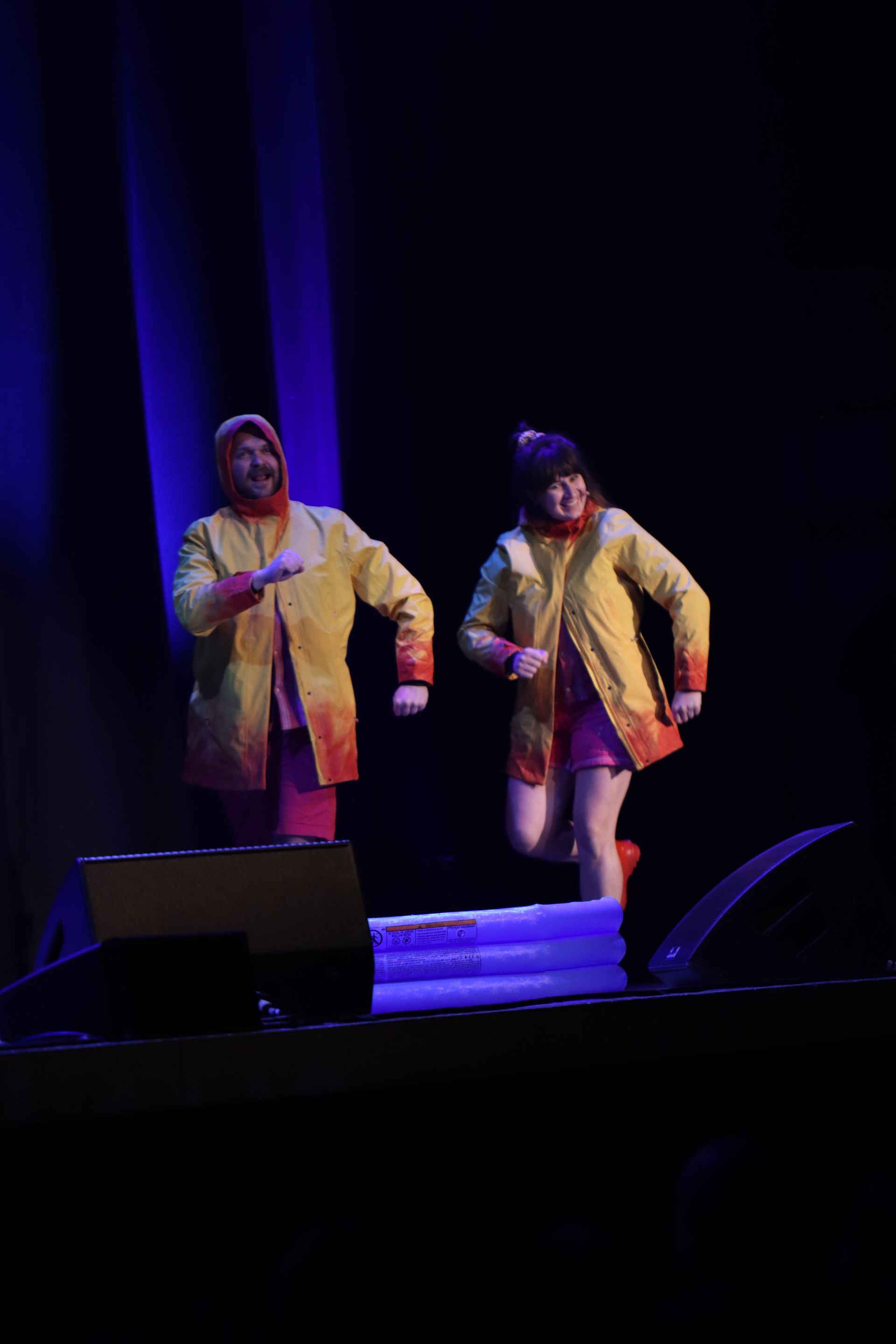 Two people in yellow raincoats and hoods onstage, running.