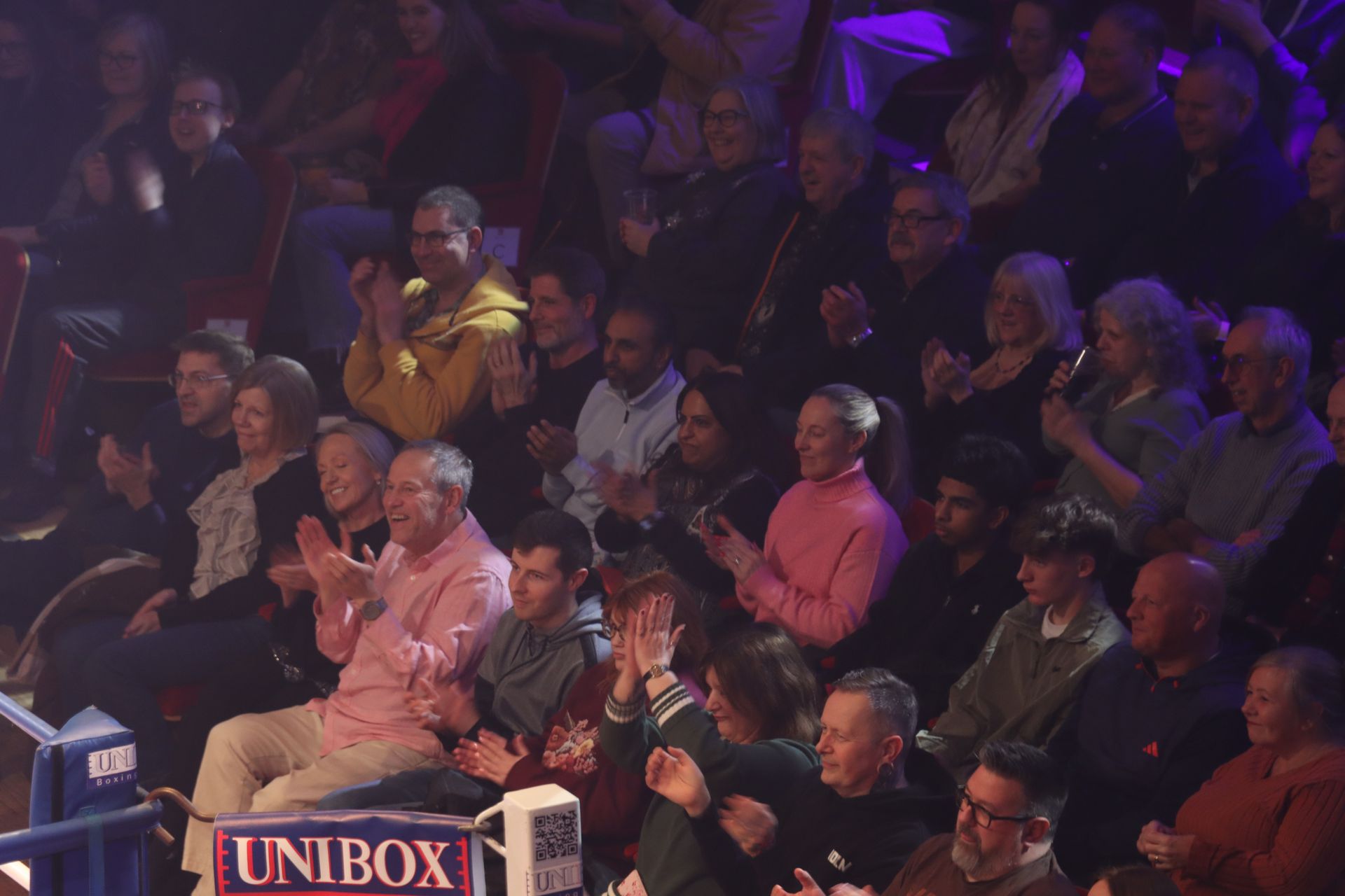 Audience applauding at an event. People in rows, illuminated by stage lights.
