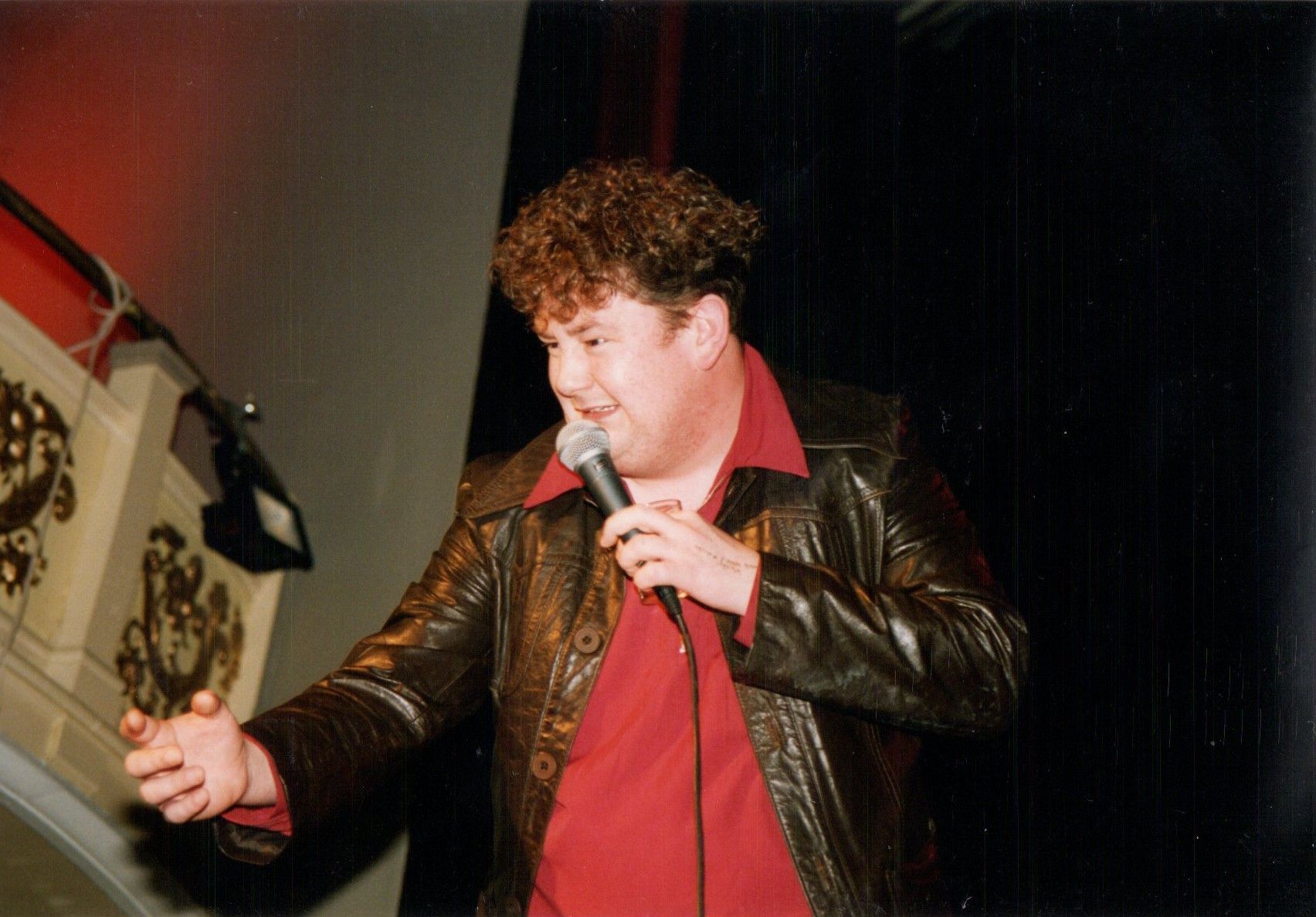 Man with curly hair in a red shirt and brown jacket holds a microphone, gesturing on stage.