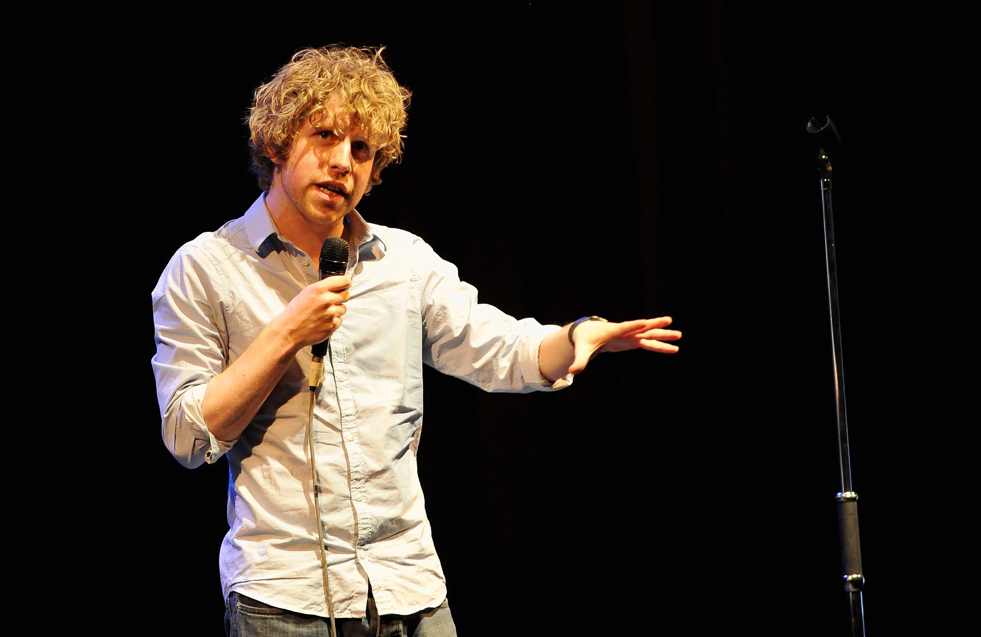 Man with curly hair, in a light blue shirt, speaks into a microphone on a stage.