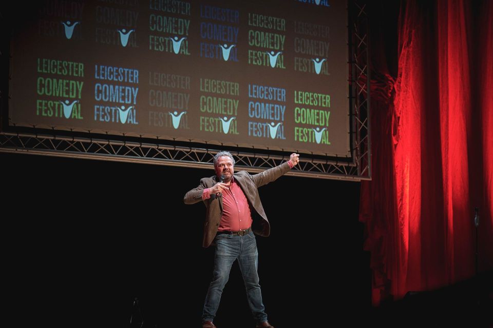 Man with long brown hair, crossed arms, looking surprised, against a blue backdrop.