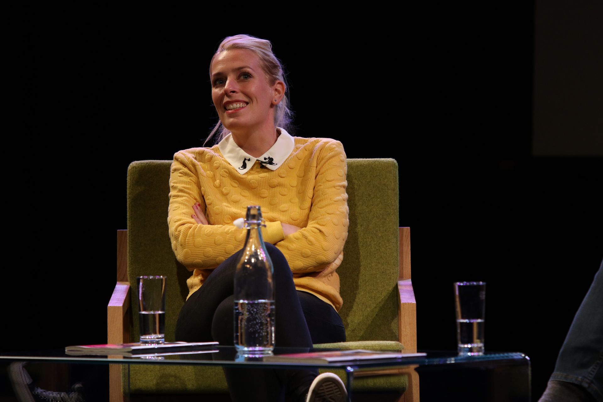Woman with blonde hair wearing a yellow sweater and white collar sits on a stage with arms crossed.