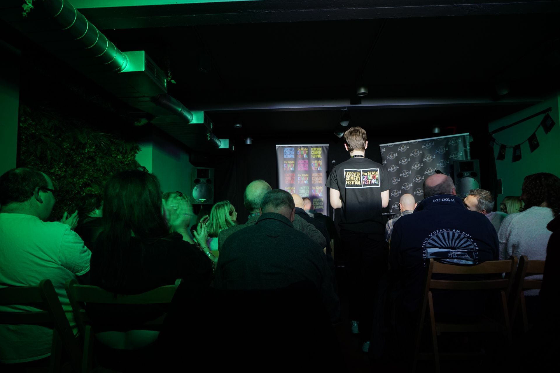 A person on stage faces a seated audience in a dimly lit room with green lighting. People clap.