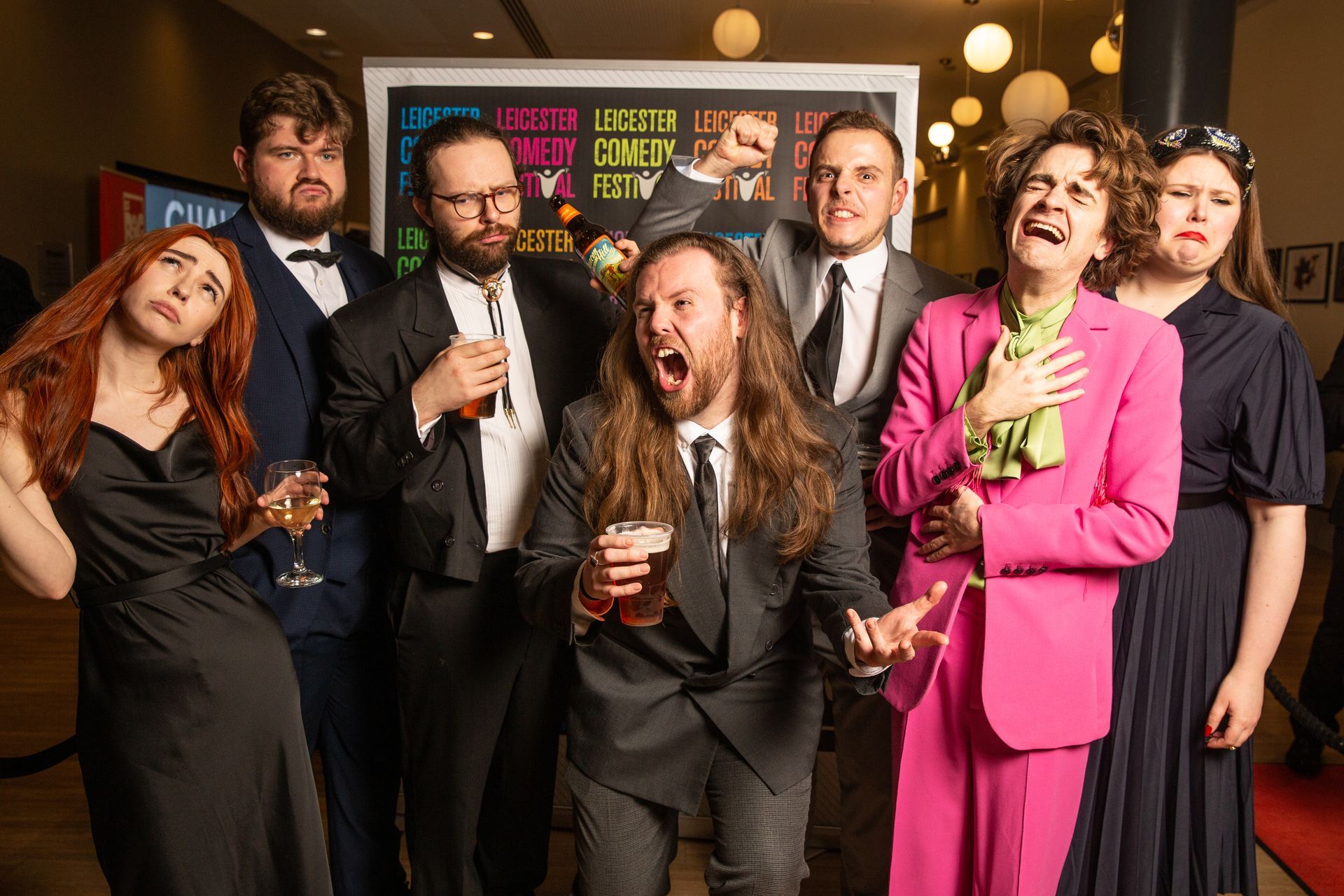 Group of people, mostly in suits, pose at a festival. Some hold drinks, with various expressions.