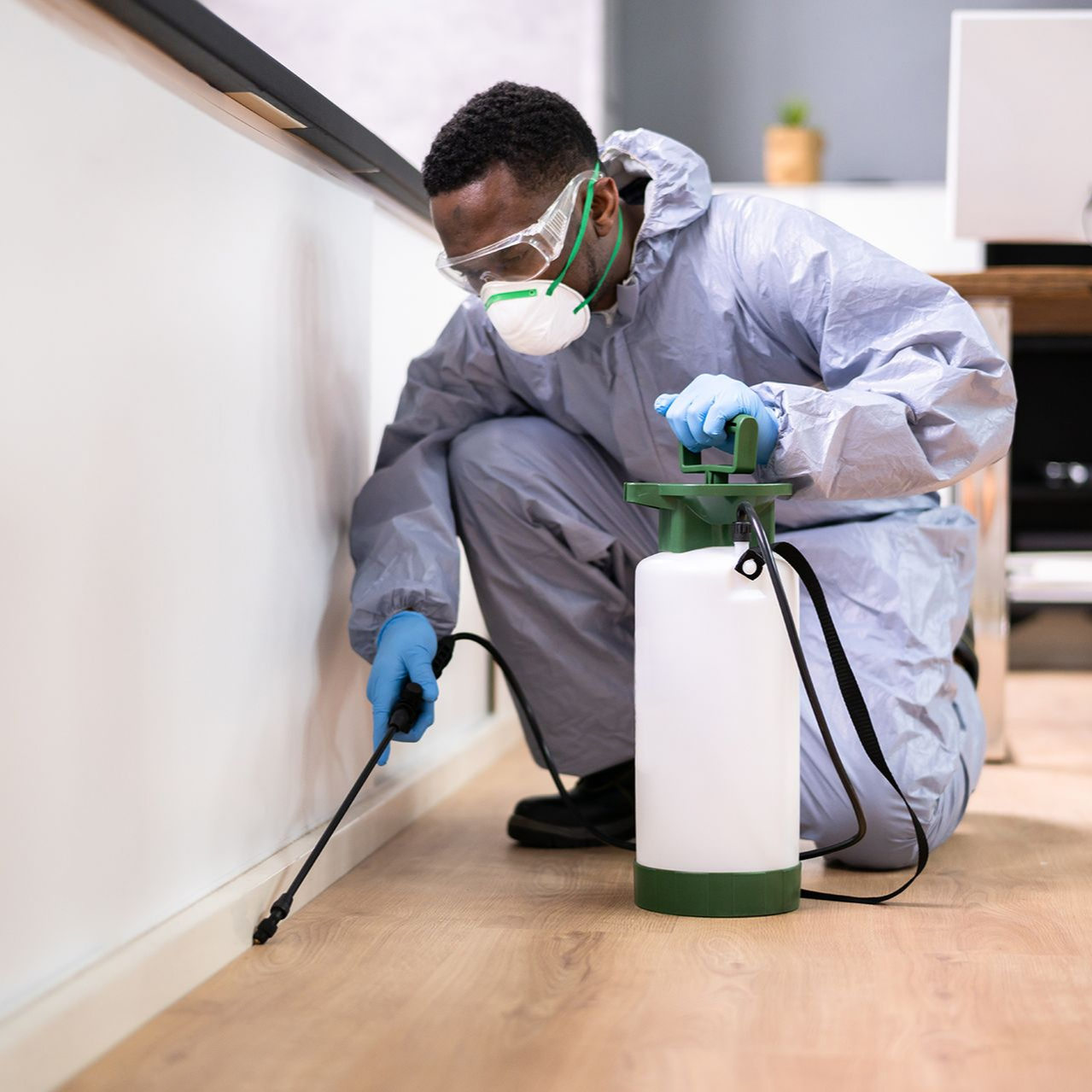 A person in a protective suit, mask, and gloves kneeling to spray pesticide along a baseboard in an indoor setting.