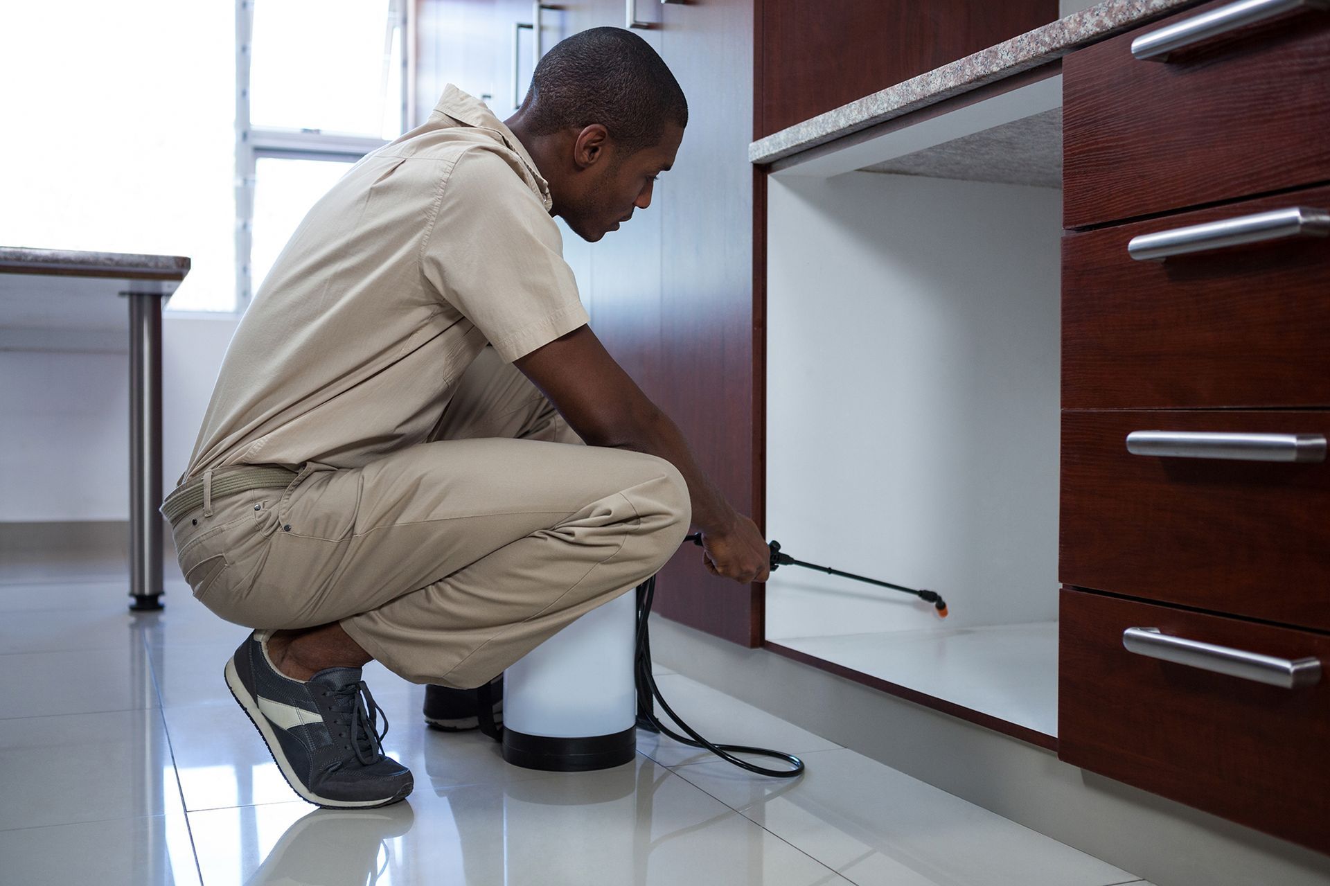 Man in beige jumpsuit spraying insecticide in a kitchen cabinet.