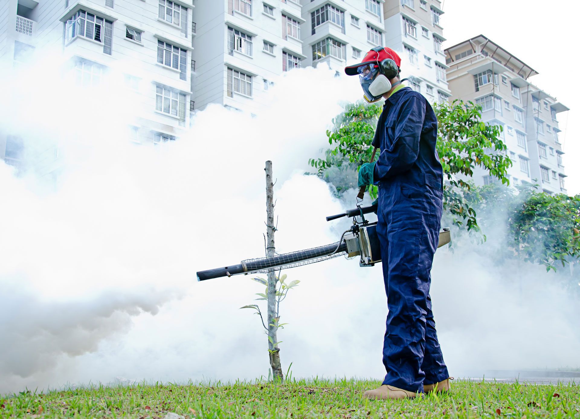 A worker in protective clothing and a respirator uses a fogging machine to spray insecticide near a residential building.
