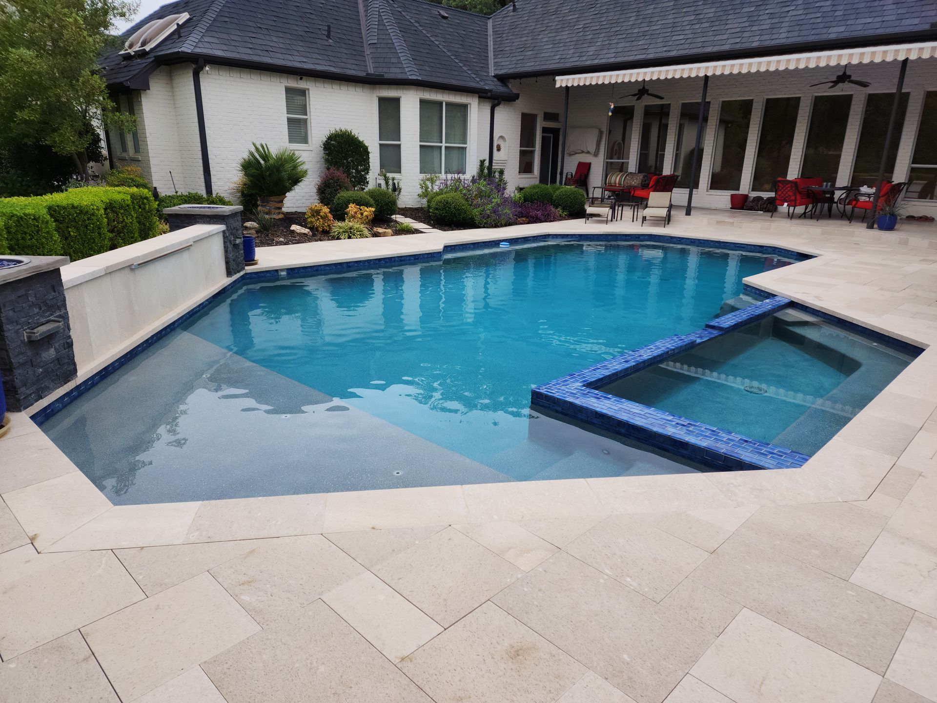 A backyard pool with a built-in spa, blue water, and light-colored stone patio, set behind a house with a covered patio.